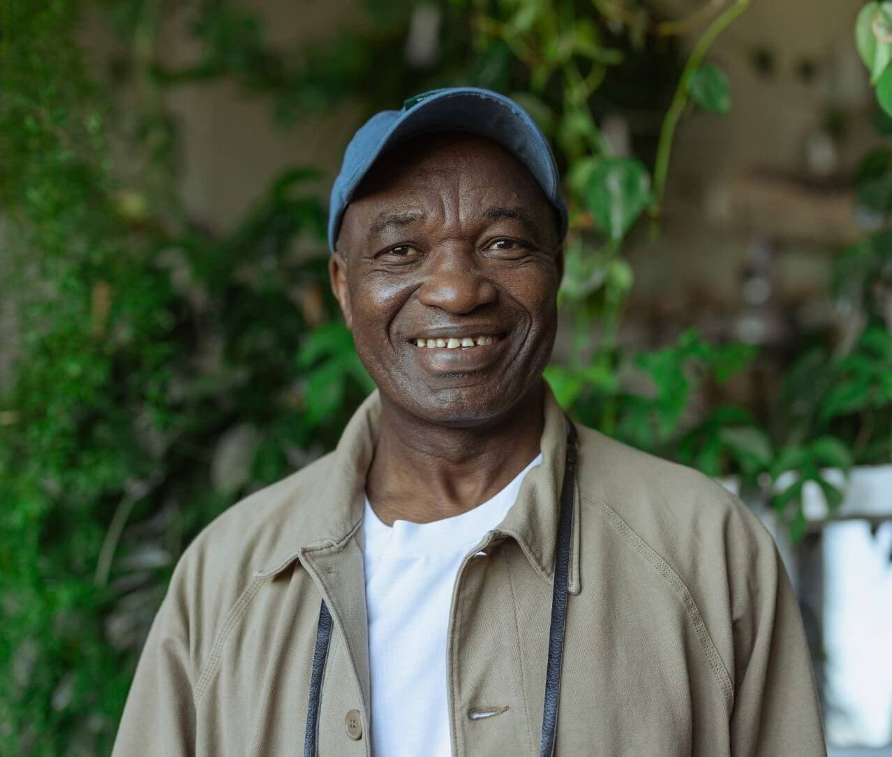 Man in a blue cap and beige jacket smiling with a garden background. - Home Instead