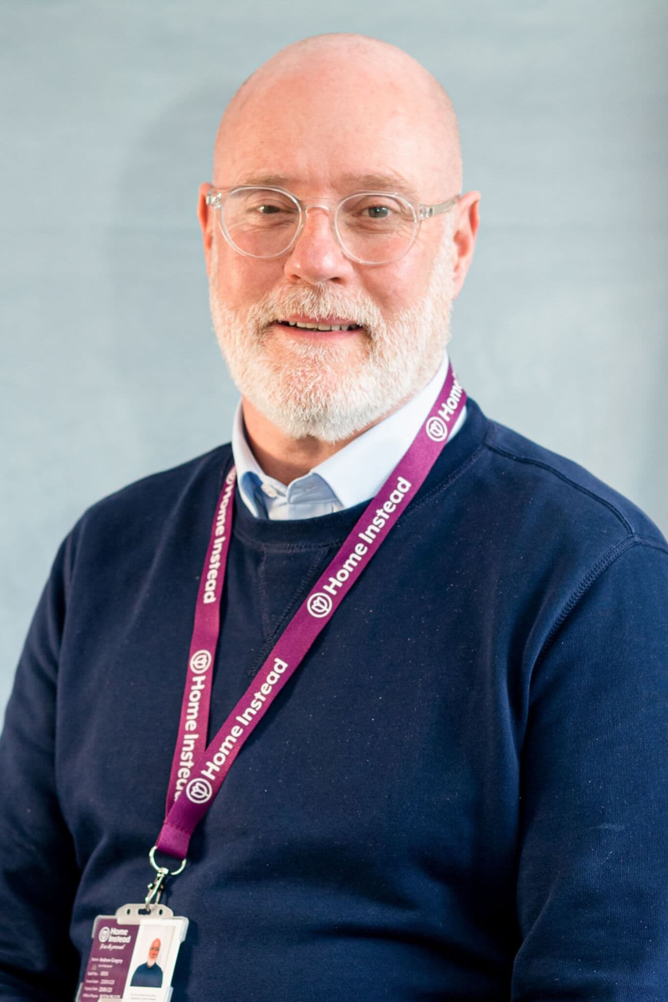 Smiling man with glasses and a beard, wearing a purple lanyard and navy blue sweater, standing against a plain background. - Home Instead