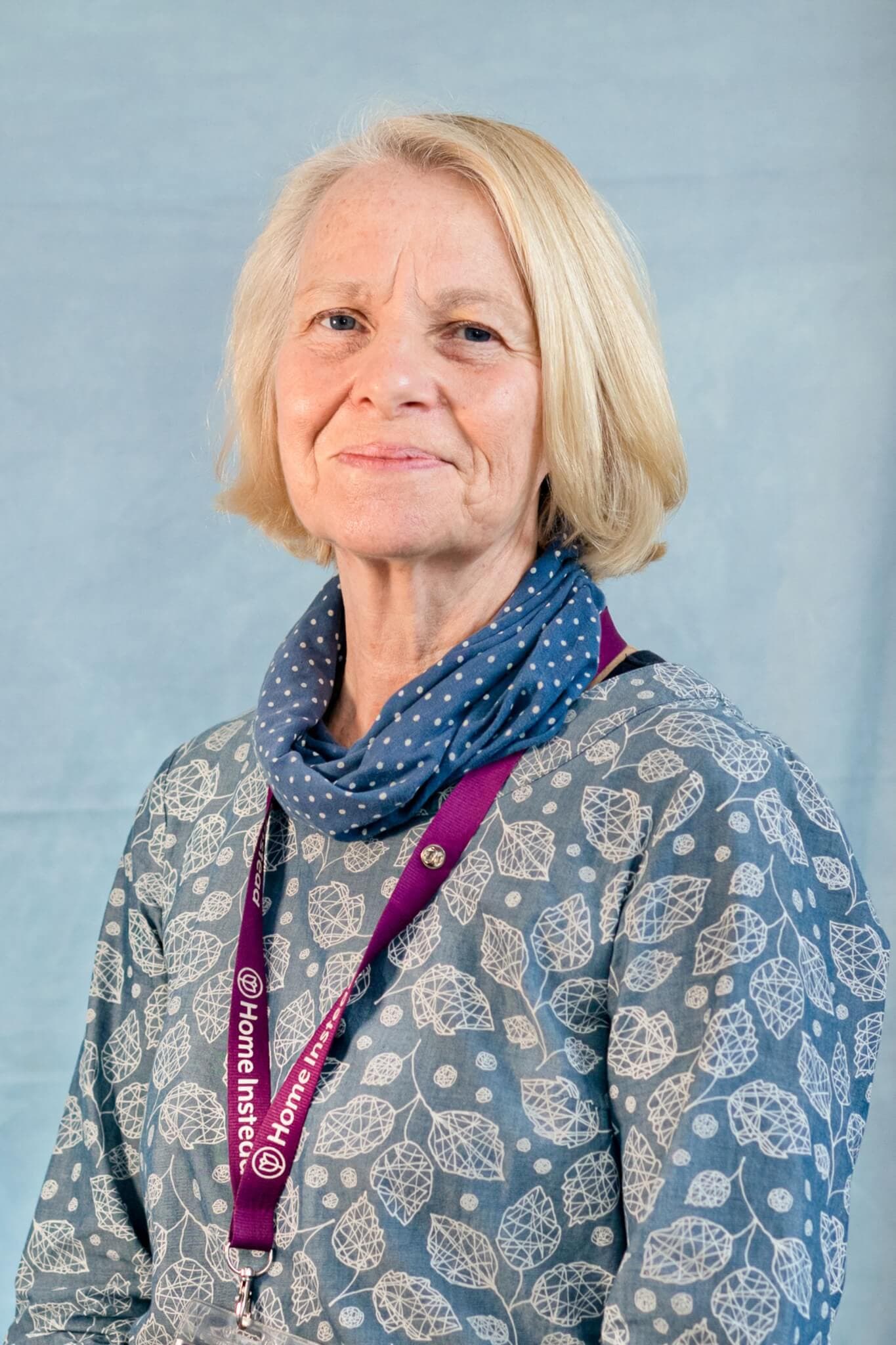 An older woman with shoulder-length blond hair and a lanyard, wearing a blue patterned top and a polka dot scarf. - Home Instead