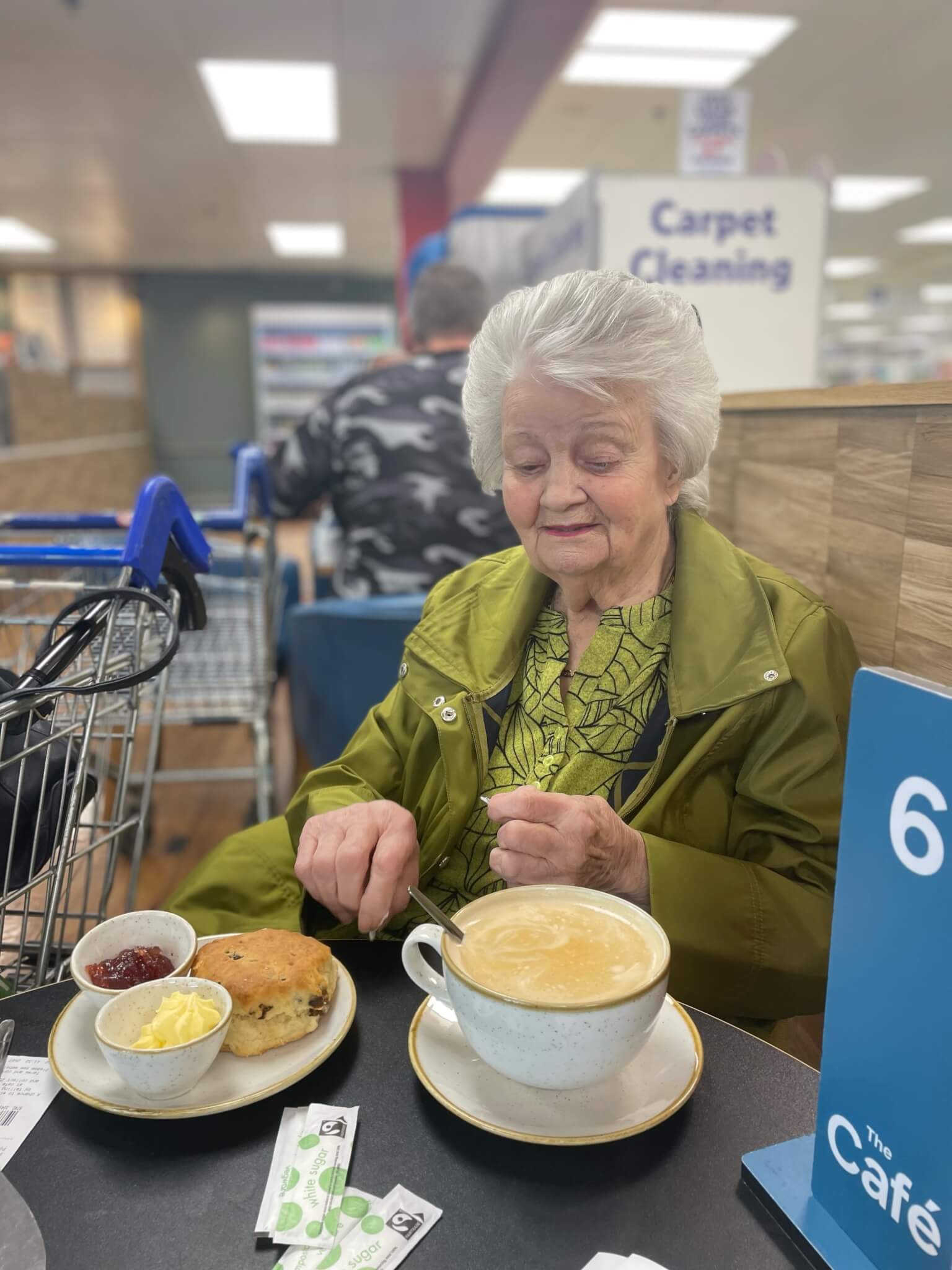 An elderly woman enjoys a scone and coffee at a cafe inside a store, with a shopping cart parked beside her. - Home Instead