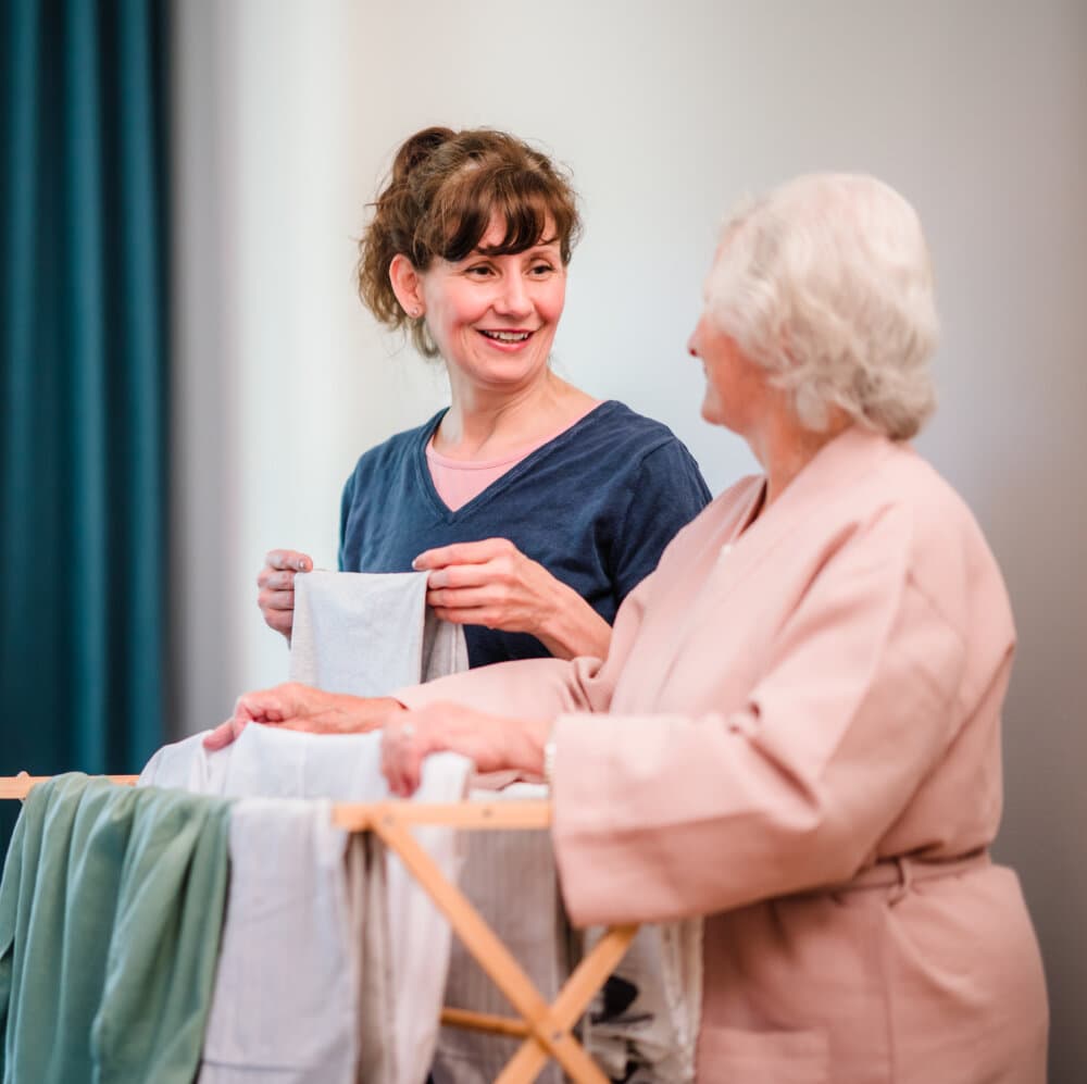 Two women smiling and chatting while hanging laundry on a drying rack indoors. - Home Instead