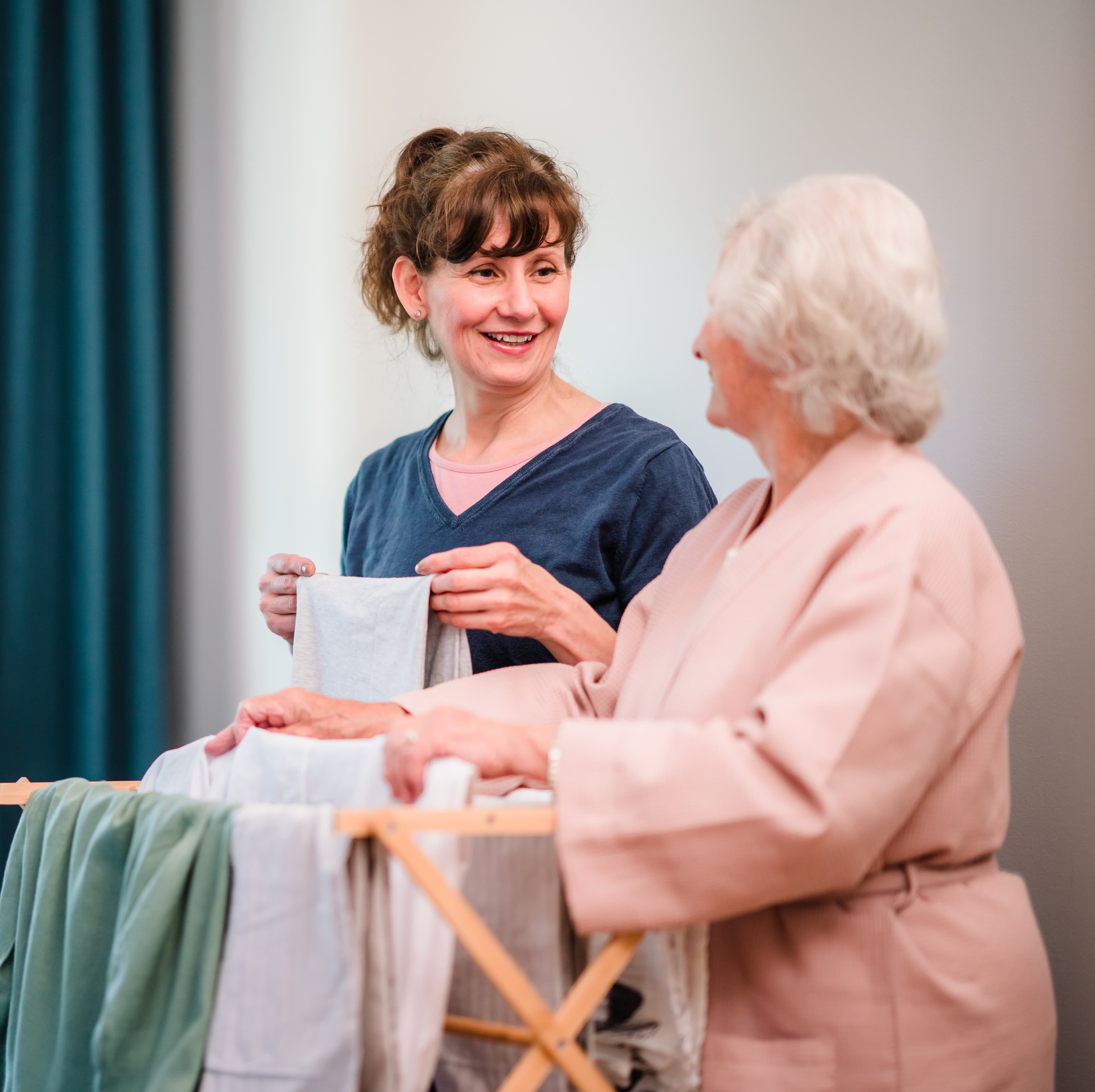 Two women smiling and chatting while hanging laundry on a drying rack indoors. - Home Instead
