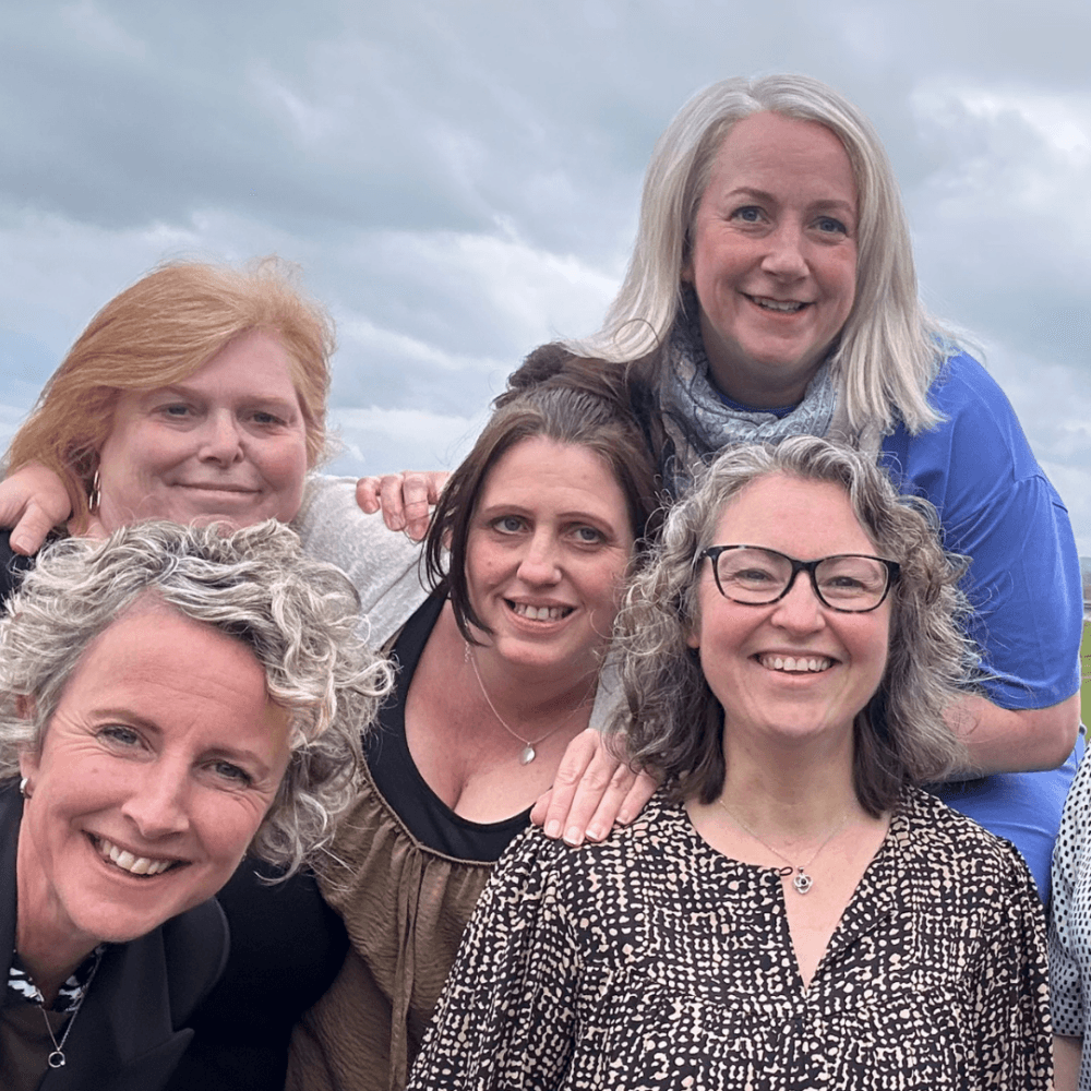 Five smiling women pose together outdoors with a cloudy sky in the background. - Home Instead
