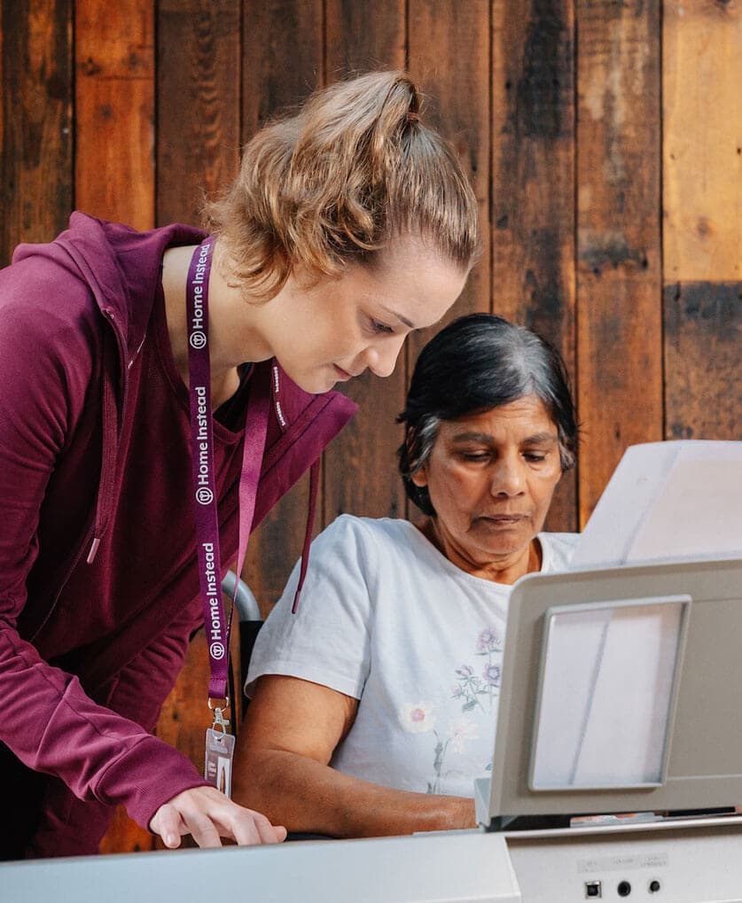 A caregiver assists an elderly woman with an activity at a table in a cozy, rustic room. - Home Instead