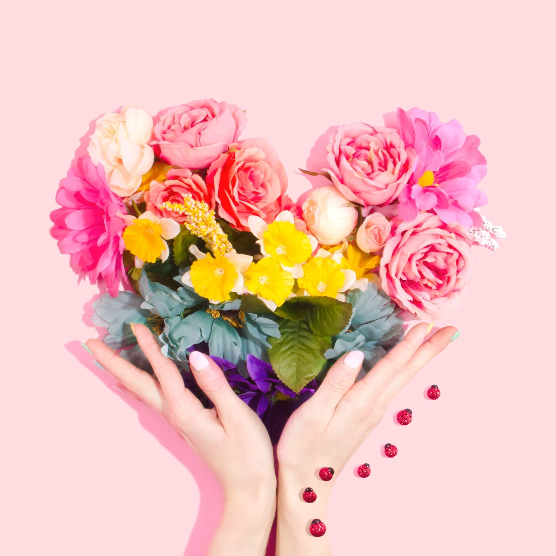 Hands holding a heart-shaped bouquet of colorful flowers on a pink background, with ladybugs nearby. - Home Instead