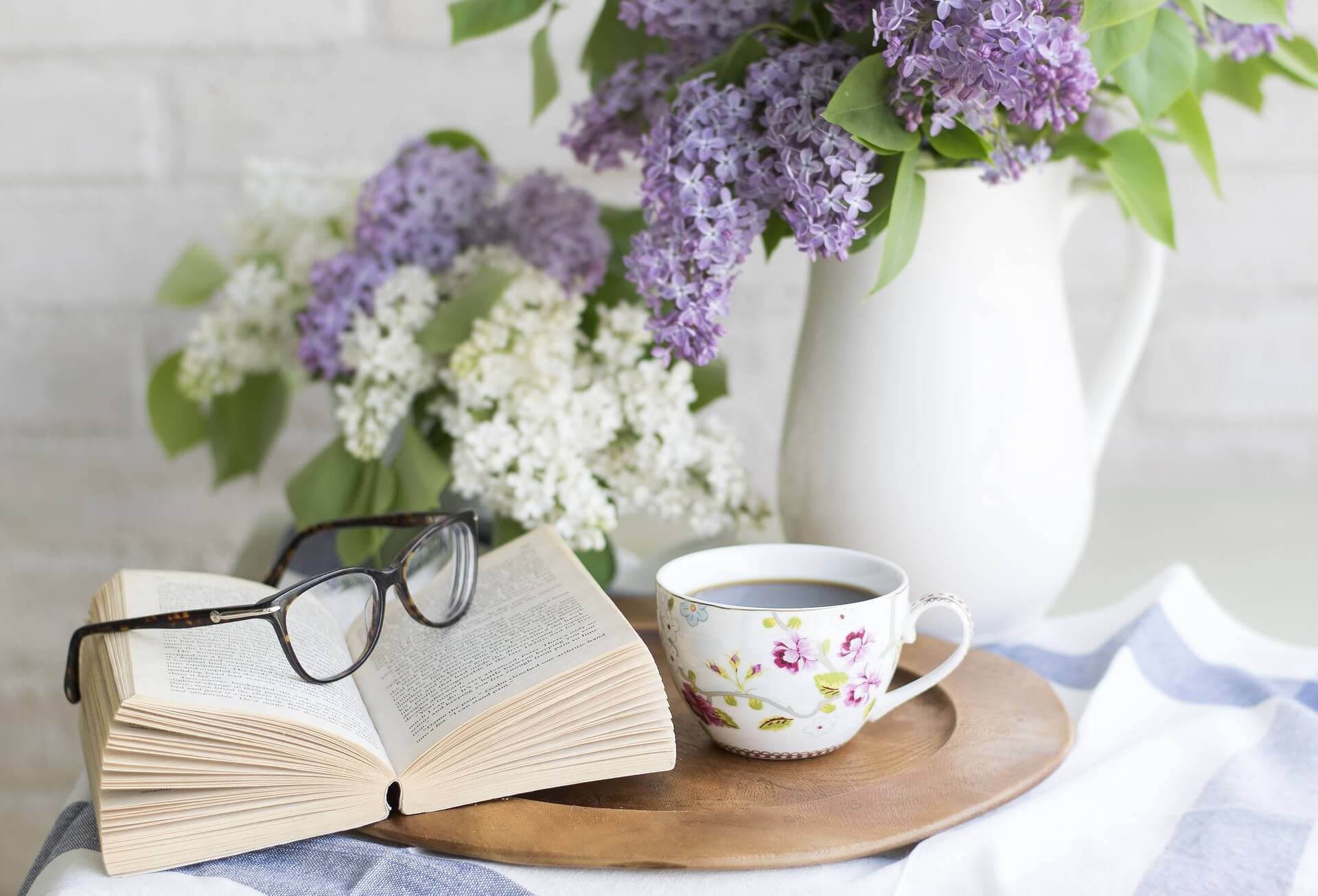 Open book with glasses, a floral teacup on a wooden tray, and a vase of lilac flowers on a white surface. - Home Instead