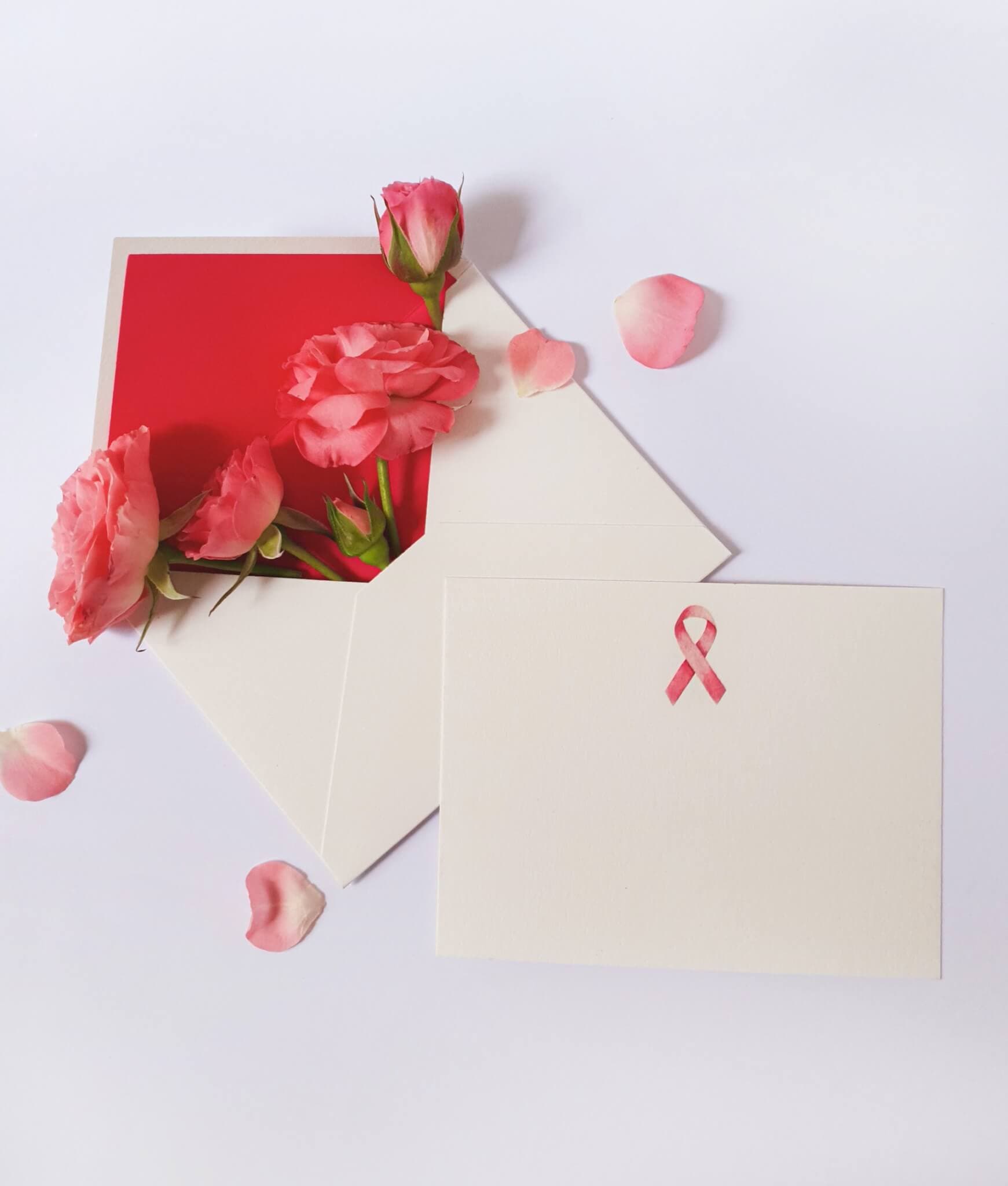 An open envelope with pink flowers and petals, next to a card featuring a pink ribbon, on a white background. - Home Instead