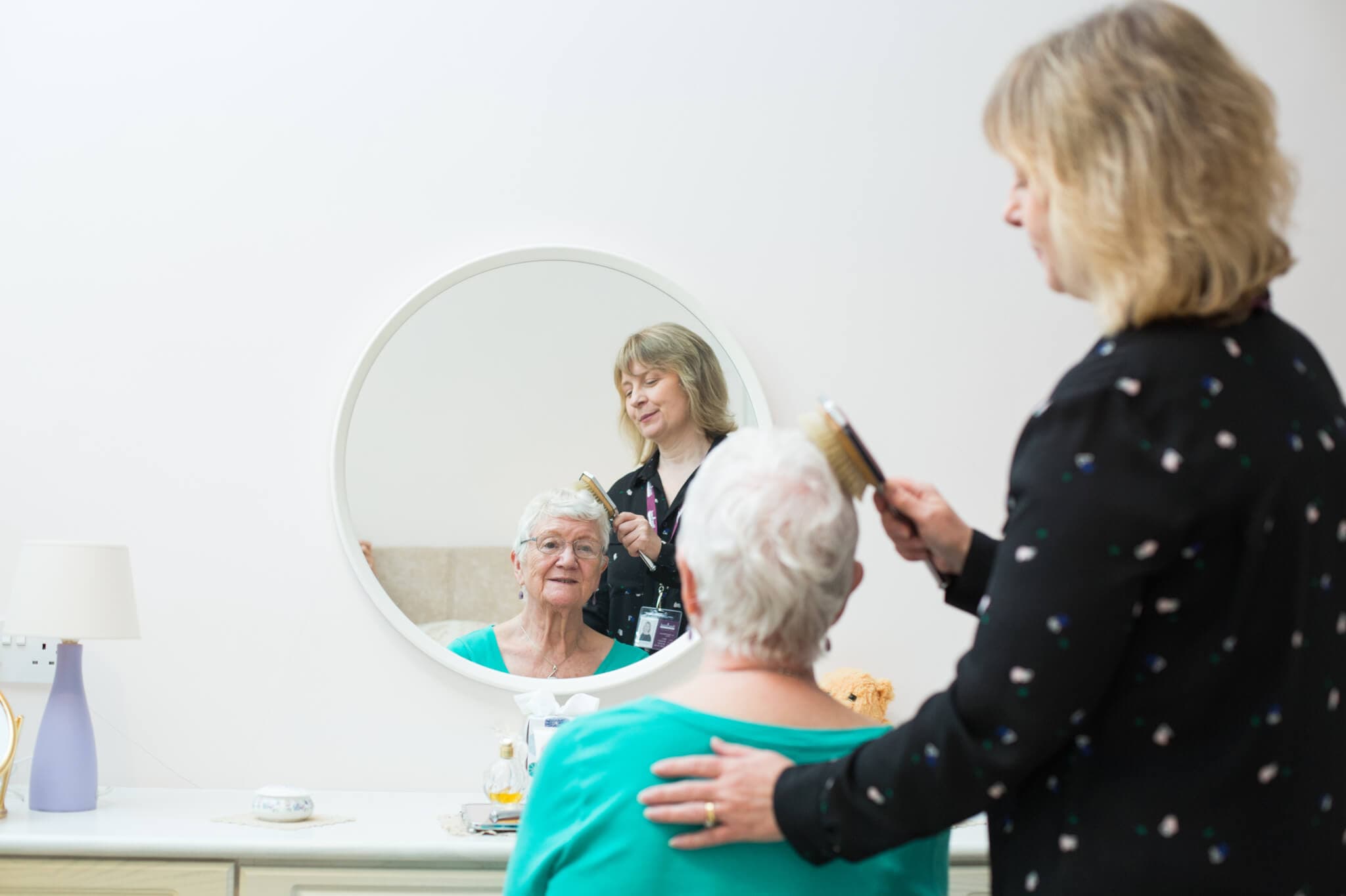 A woman brushes the hair of an elderly woman sitting in front of a mirror, both reflected in the mirror's view. - Home Instead