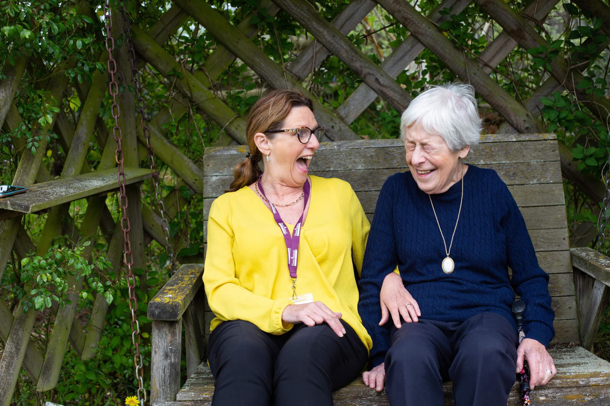 Two women sitting on a wooden bench, laughing and enjoying a moment together against a leafy background. - Home Instead