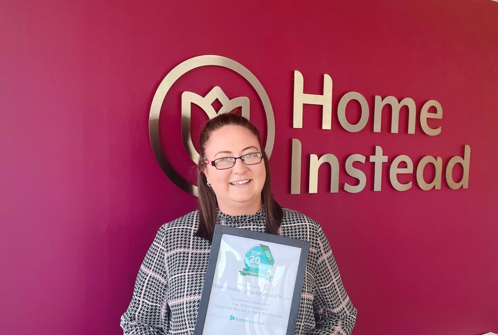 A person holding a framed certificate stands in front of a burgundy wall with the Home Instead logo and sign. - Home Instead