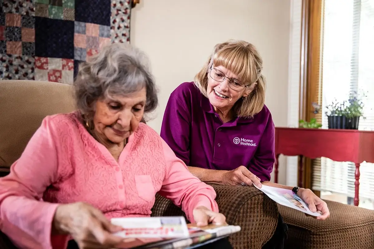 An elderly woman reads a book while a caregiver in a purple Home Instead shirt smiles and talks to her. - Home Instead