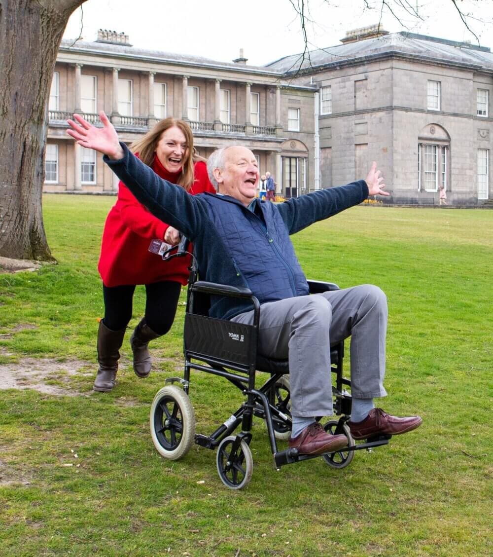 Home Instead client spreading his arms joyfully as his Home Instead carer pushes him downhill in the grounds of a stately home.