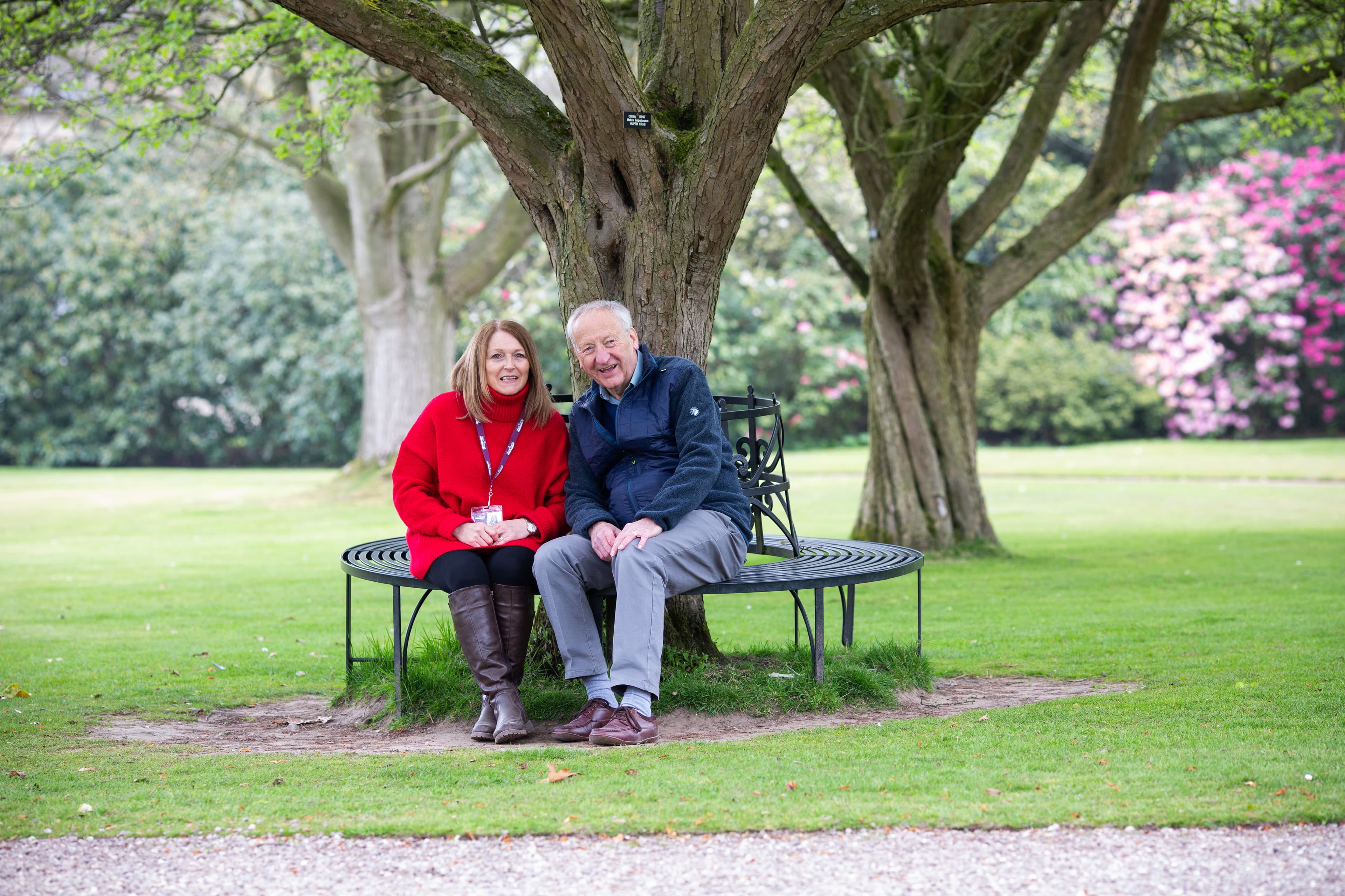 An elderly couple sitting on a circular bench under a large tree in a garden, smiling and enjoying the day. - Home Instead