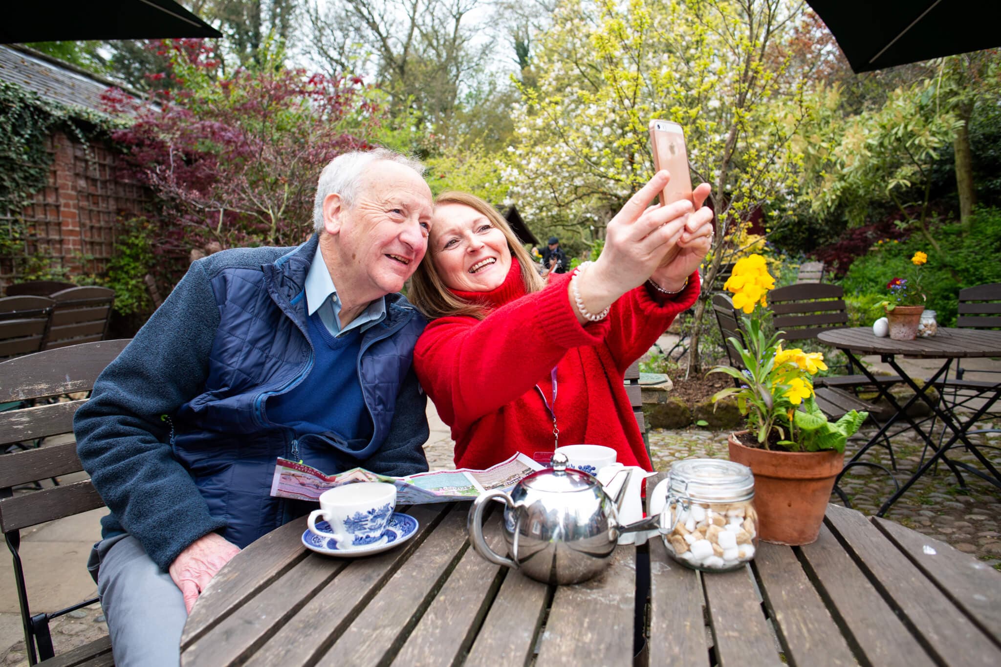 A senior man and woman smile while taking a selfie at an outdoor café table with tea and flowers. - Home Instead