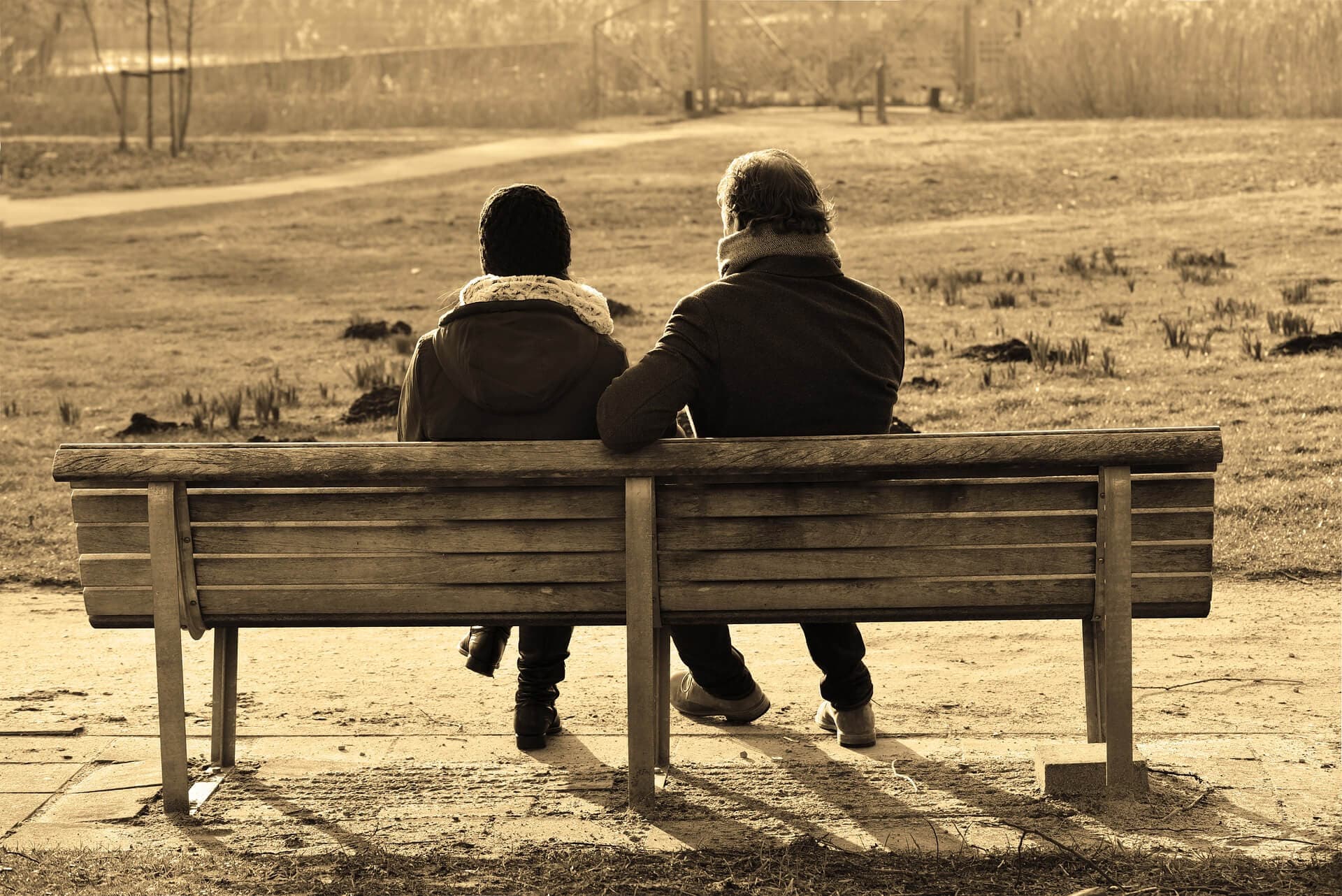 Two people sitting on a wooden bench in a park, viewed from behind, with a serene and peaceful setting. - Home Instead