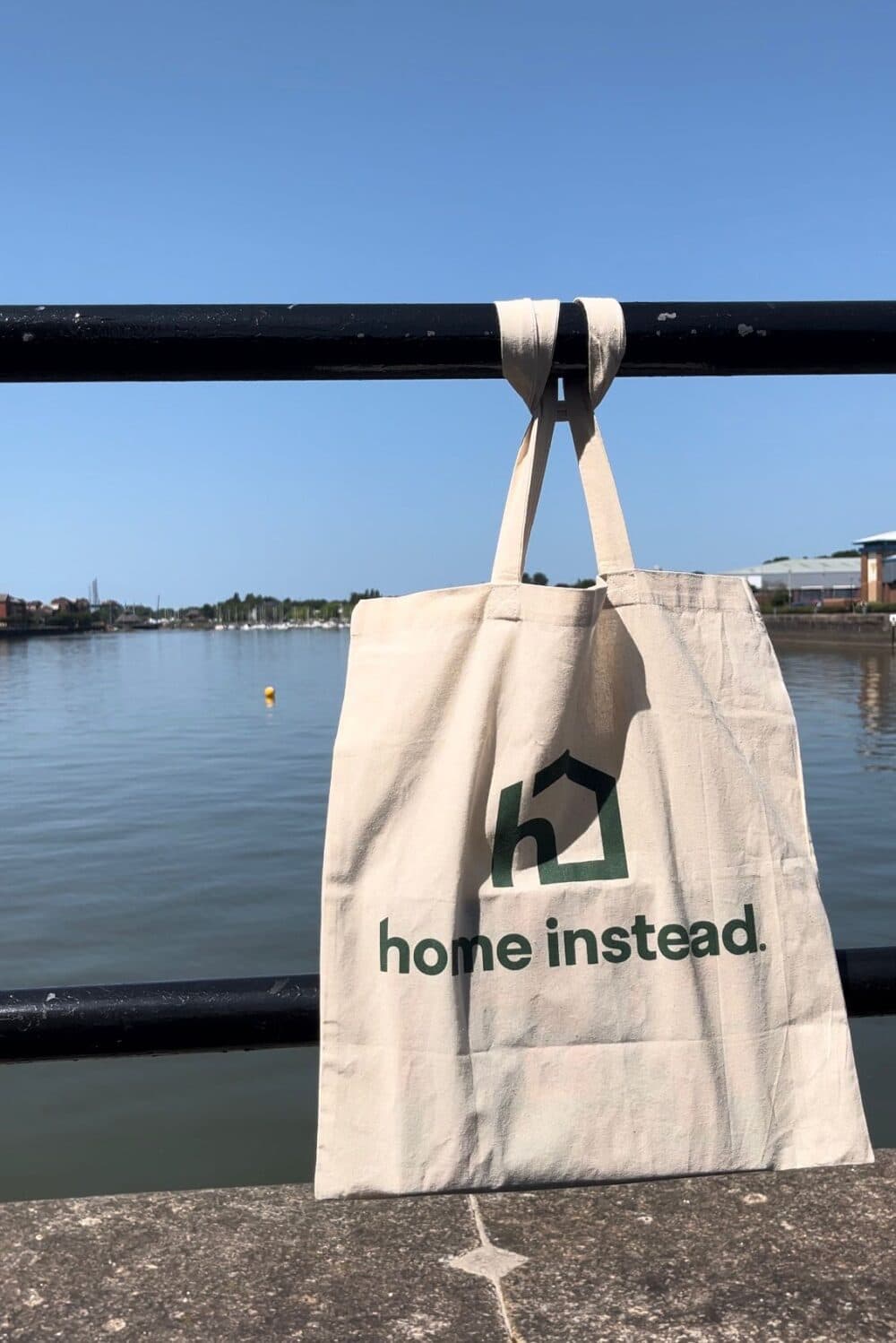 Beige tote bag with "home instead" logo hanging on a railing by a waterfront under a clear sky. - Home Instead