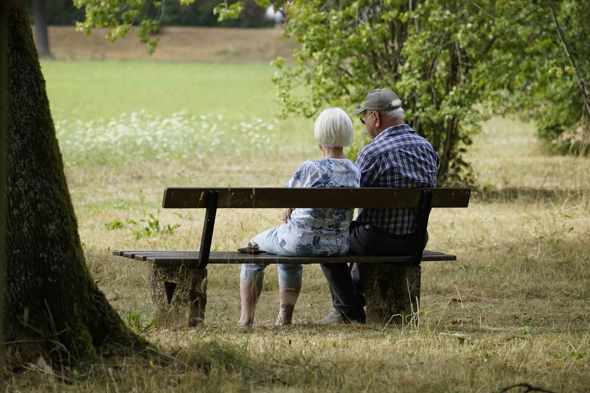 A senior couple sitting on a bench in a park, surrounded by trees and grass, facing away from the camera. - Home Instead