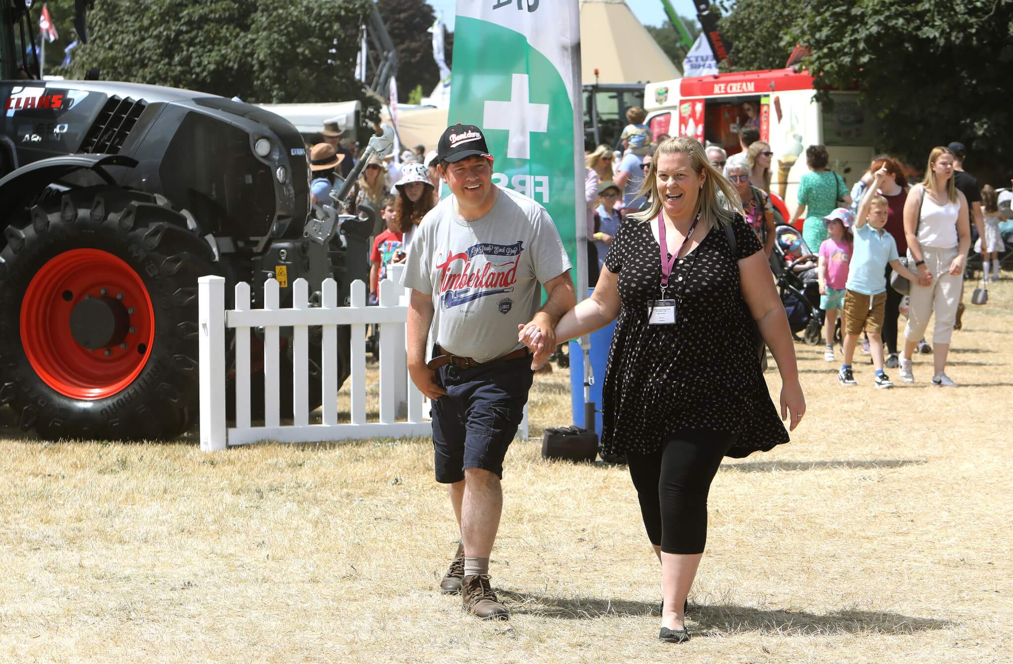 A man and a woman walk hand-in-hand at an outdoor event with a tractor and a crowd in the background. - Home Instead
