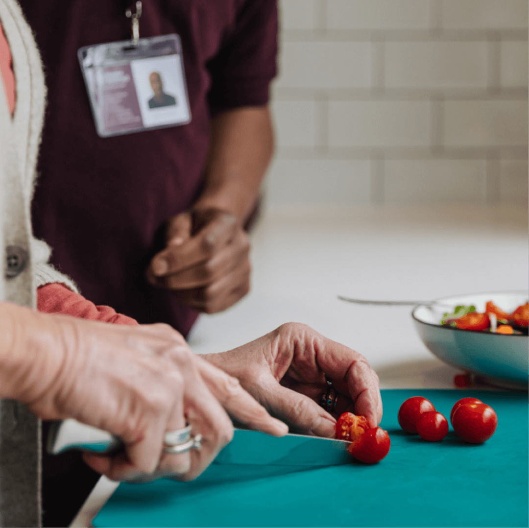 A home care client is guided by their Home Instead Care Professional to cut cherry tomatoes on a blue cutting board.
