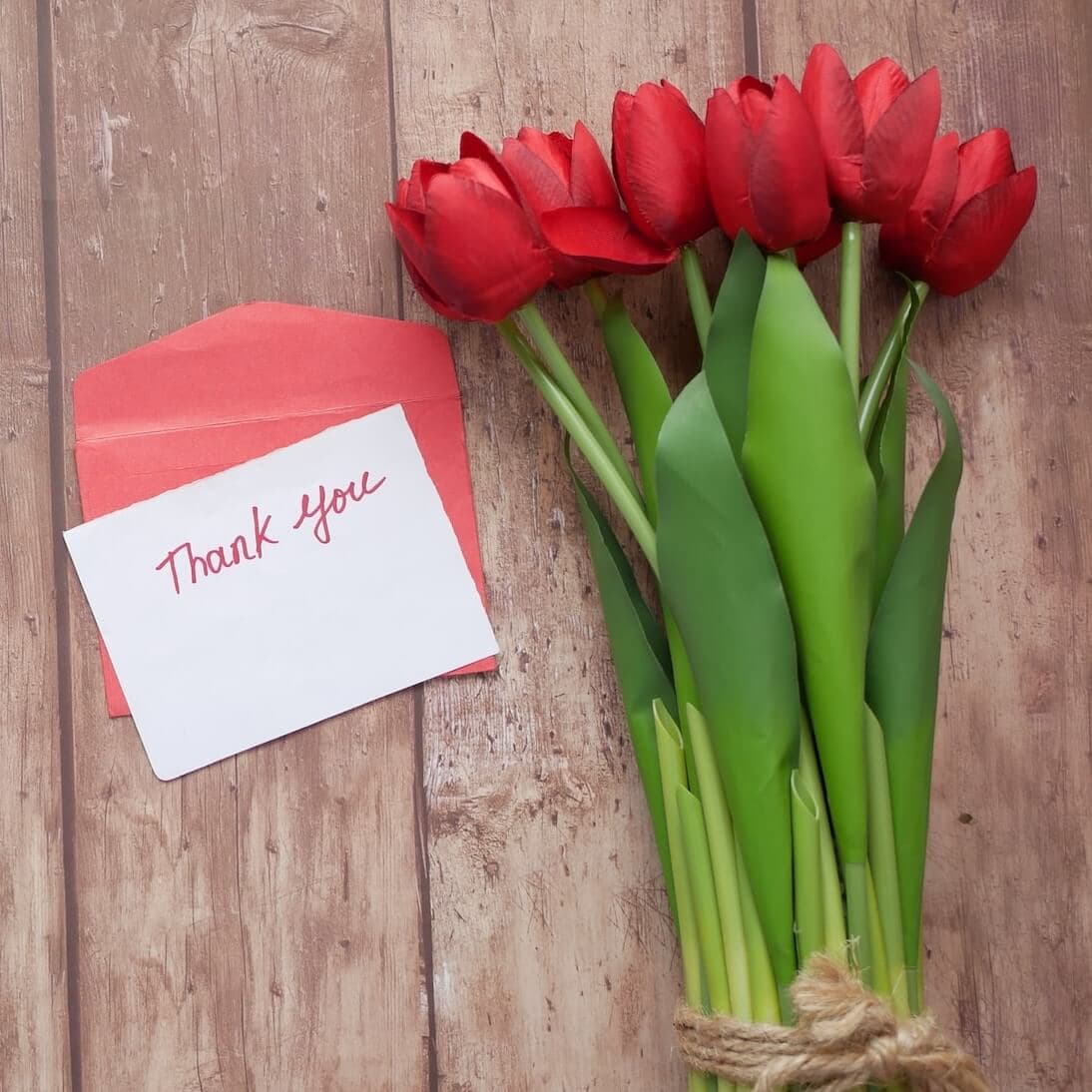 A bouquet of red tulips and a red envelope with a "Thank you" note on a wooden surface. - Home Instead