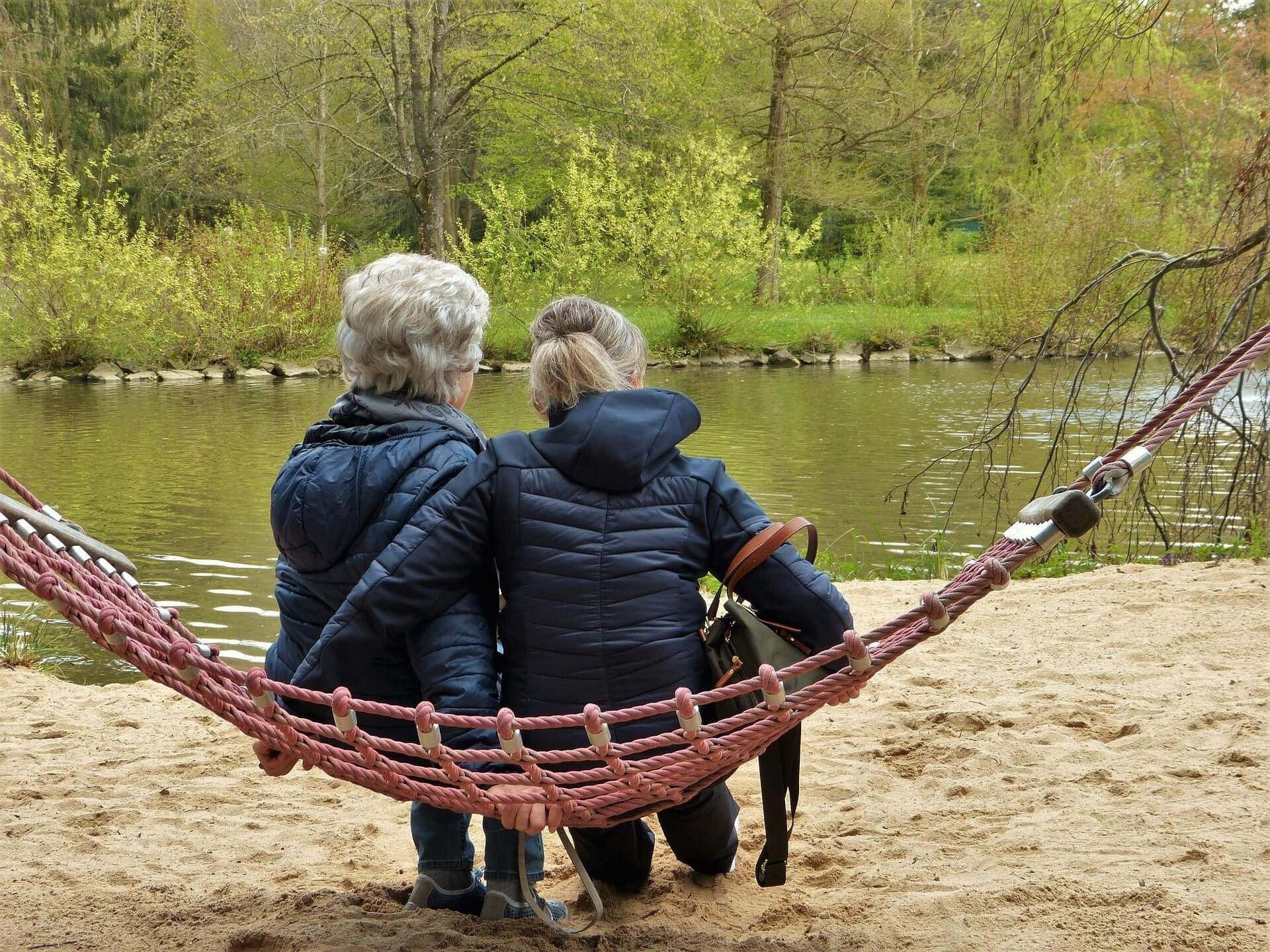 Two people wearing jackets sit on a red hammock by a riverbank, with trees and greenery in the background. - Home Instead
