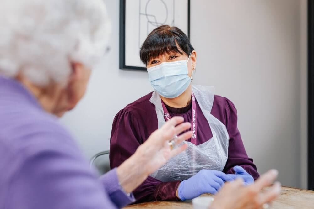 A caregiver wearing a mask and gloves talks to an elderly woman in a purple sweater in an indoor setting. - Home Instead