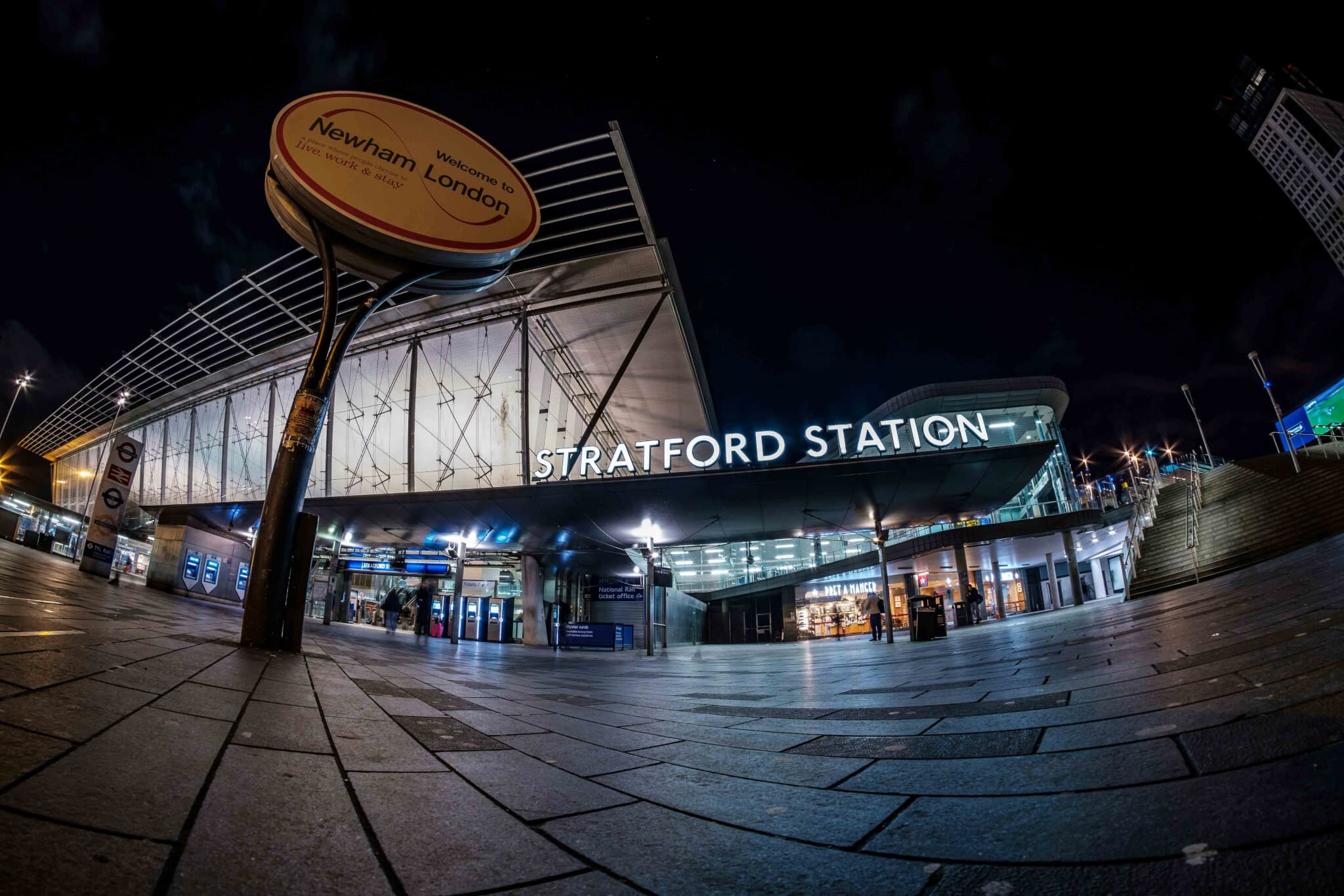 Wide-angle view of Stratford Station entrance at night with the Newham London welcome sign in the foreground. - Home Instead
