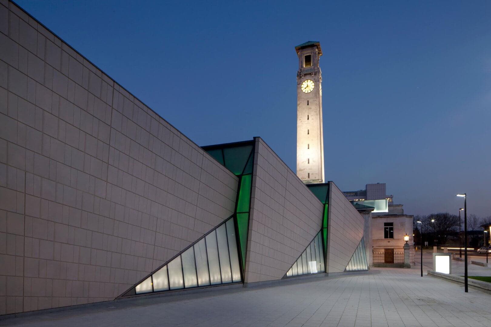Modern building with angled walls and large windows at dusk, with a tall clock tower illuminated in the background. - Home Instead Southampton