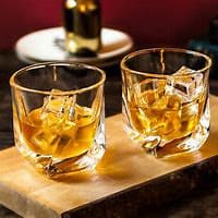 Two glasses of whiskey with ice cubes on a wooden surface, a bottle and plate in the blurred background. - Home Instead