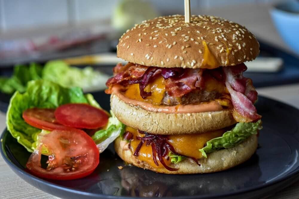 A double bacon cheeseburger with lettuce and tomato slices on a black plate, held together by a skewer. - Home Instead