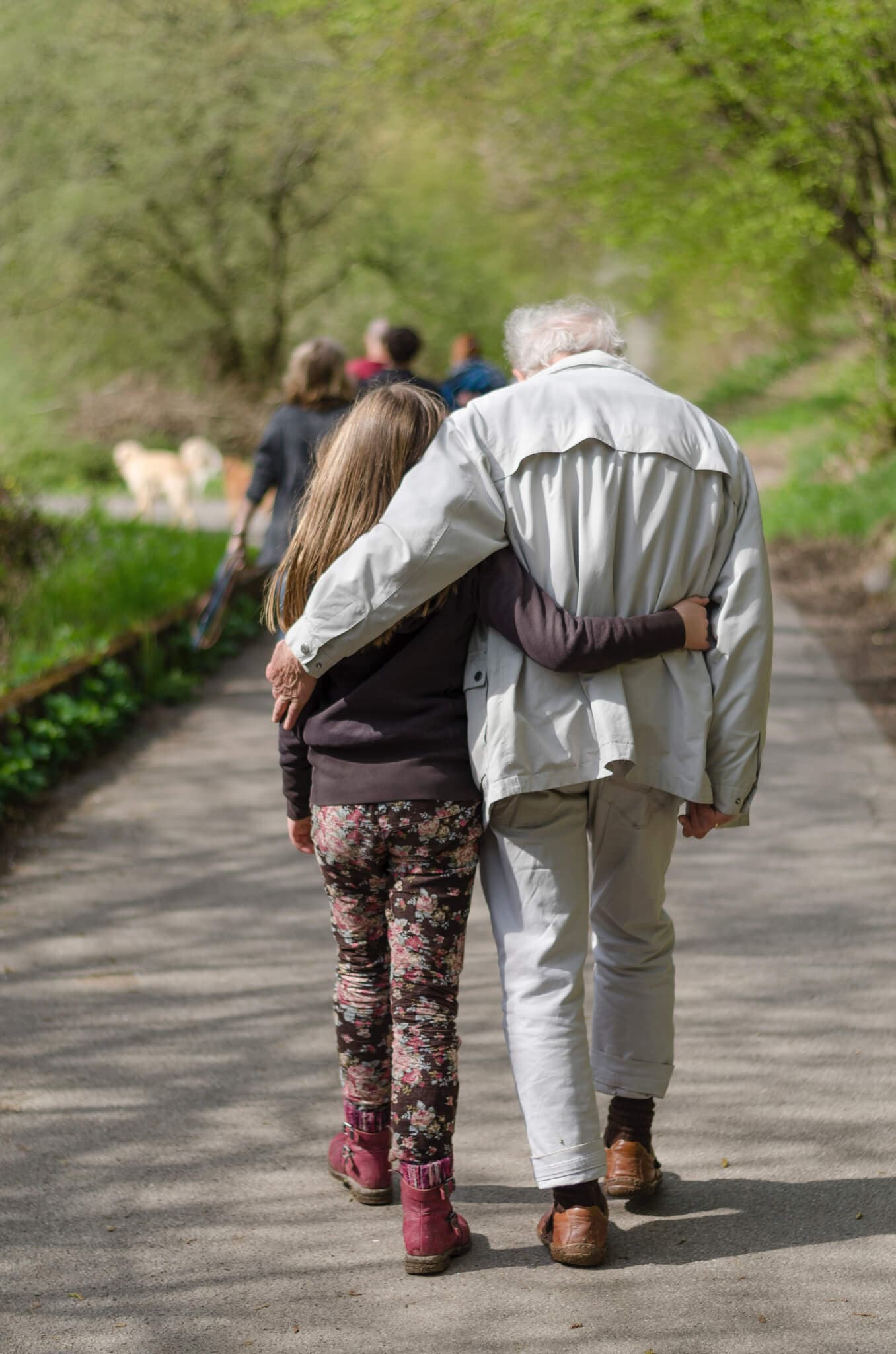 A child and an elderly person walking arm-in-arm on a forest path away from the camera. - Home Instead