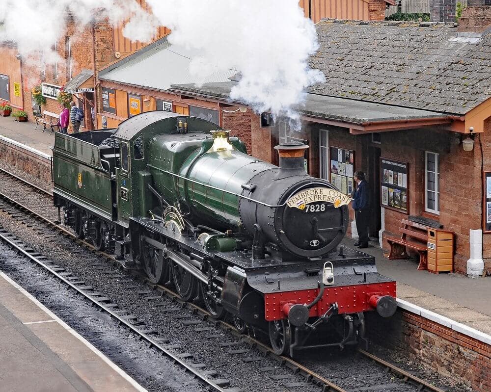 A classic steam locomotive labeled "7828" emits steam at a train station with passengers and a red brick building nearby. - Home Instead