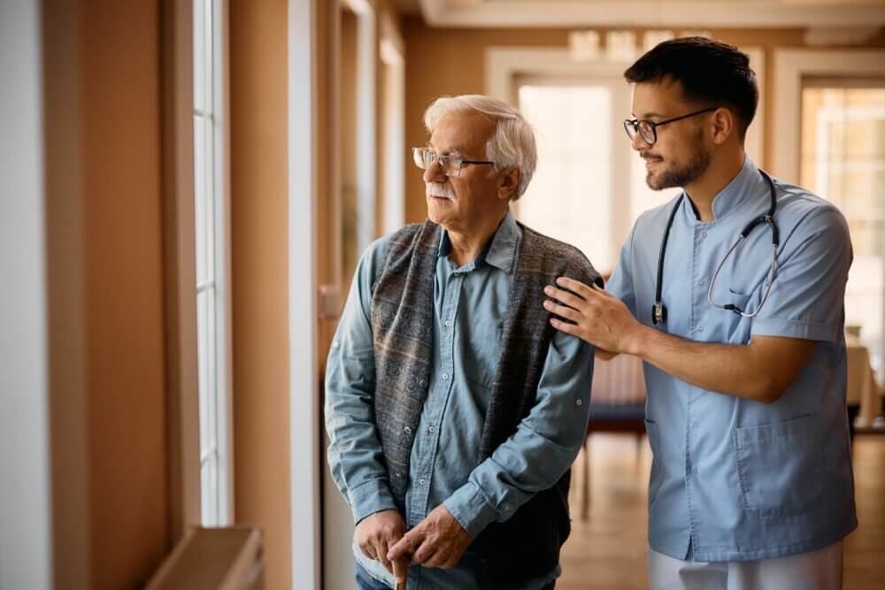 A caregiver gently supporting an elderly man with a cane as they look out of a window in a well-lit room. - Home Instead