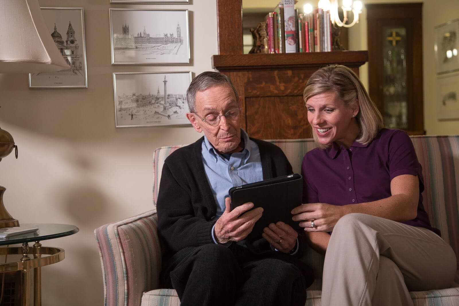 An elderly man and a woman sitting on a couch, looking at a tablet and smiling. - Home Instead
