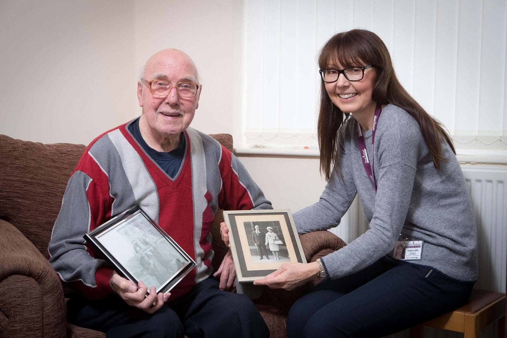 An elderly man and a woman showing framed photos, sitting on a couch in a warmly lit room. - Home Instead