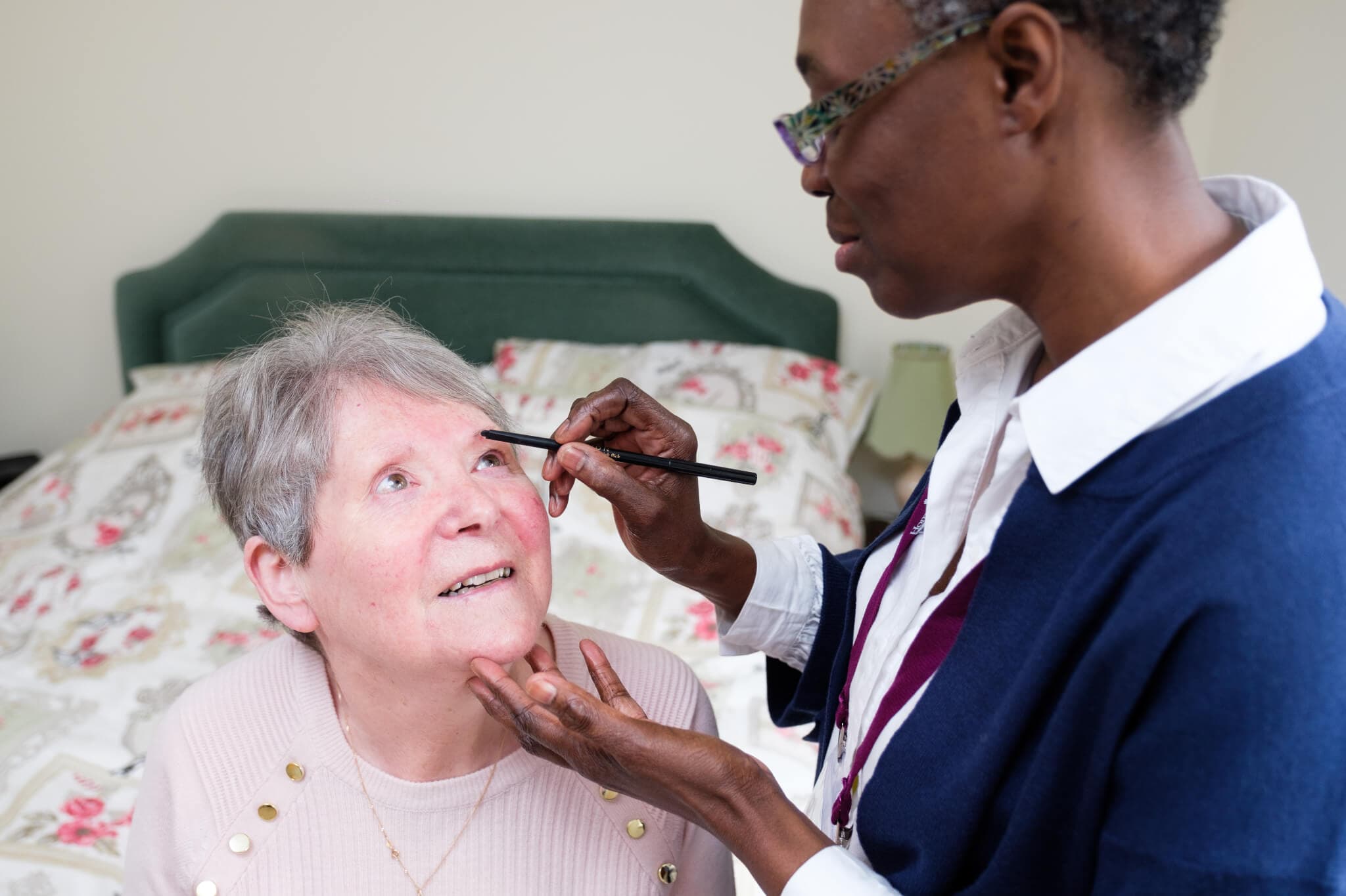 A woman applies makeup to another woman's face in a bedroom with a floral bedspread. - Home Instead
