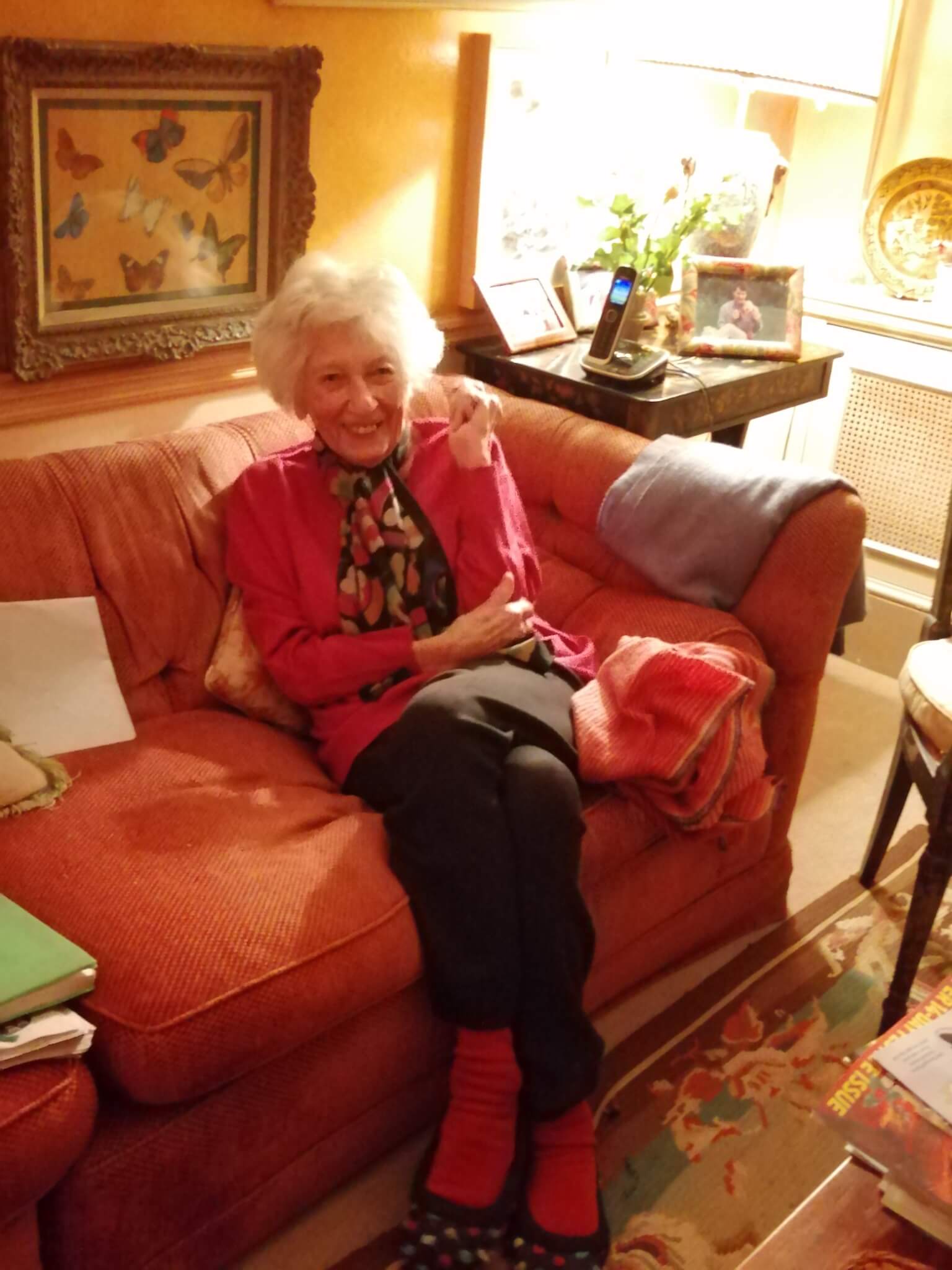 An elderly woman with white hair sits on a red couch, smiling and waving in a warmly decorated room. - Home Instead