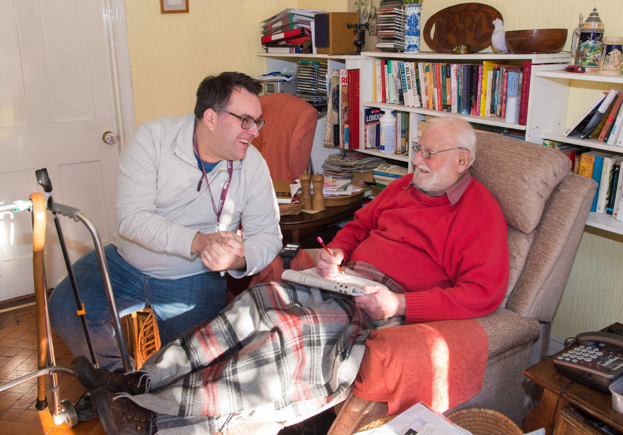 A man with glasses talks to an elderly man sitting in a chair with a blanket, surrounded by books. - Home Instead