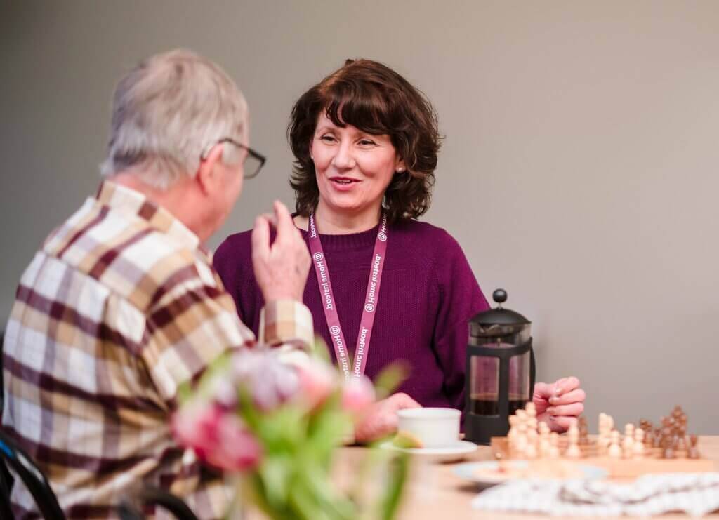 A man and a woman chatting at a table with a chess set. - Home Instead