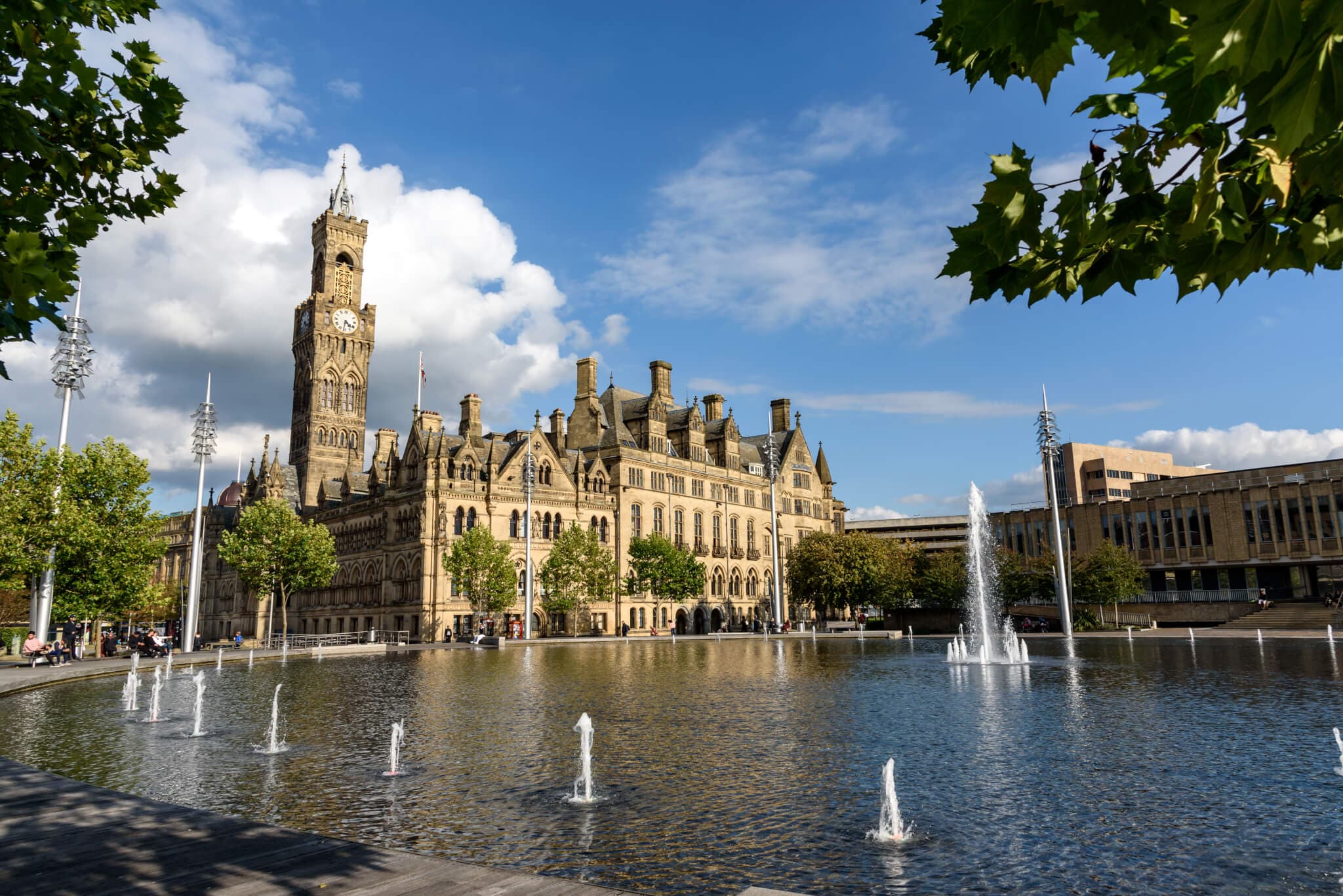 Historic building with clock tower by a fountain-filled reflective pool, surrounded by green trees on a sunny day. - Home Instead