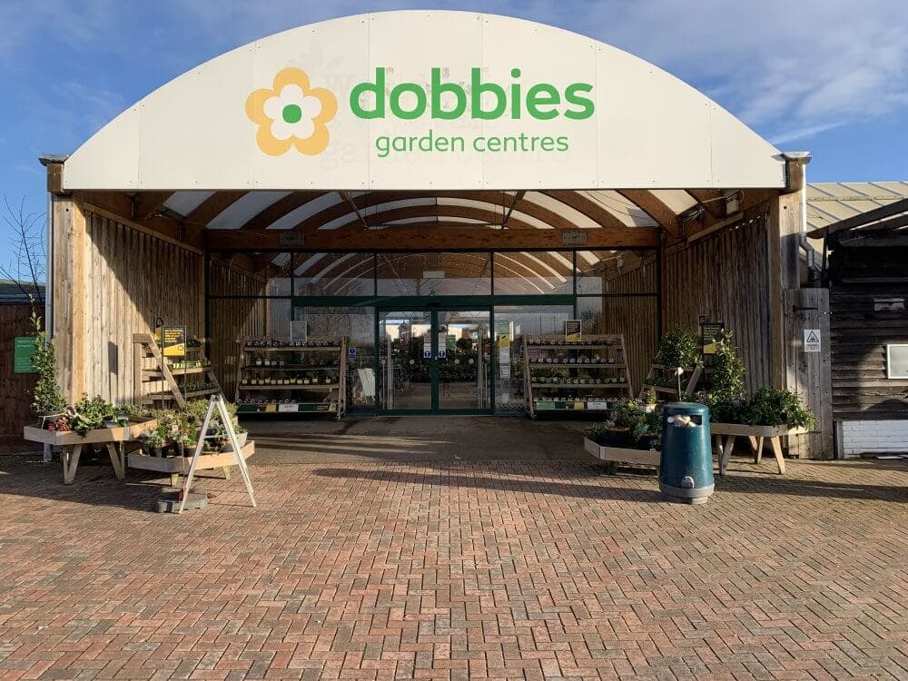 Entrance of Dobbies Garden Centre with potted plants on display, featuring a large sign above the doorway. - Home Instead
