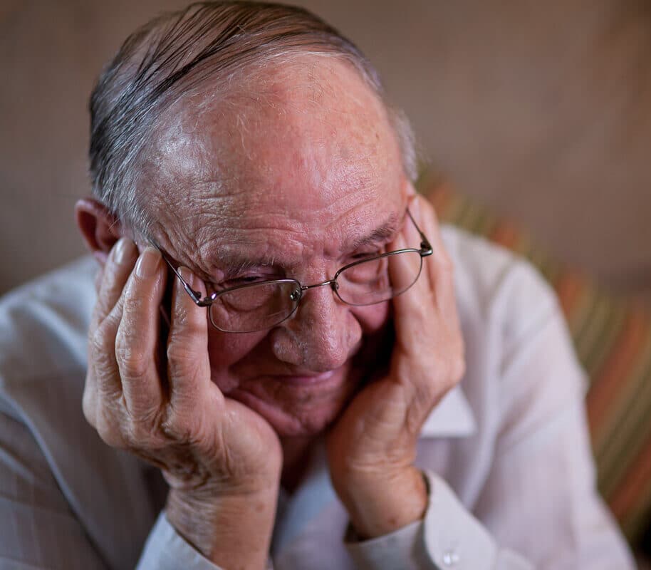 Elderly man wearing glasses, holding his head with both hands, appears to be deep in thought or feeling emotional.