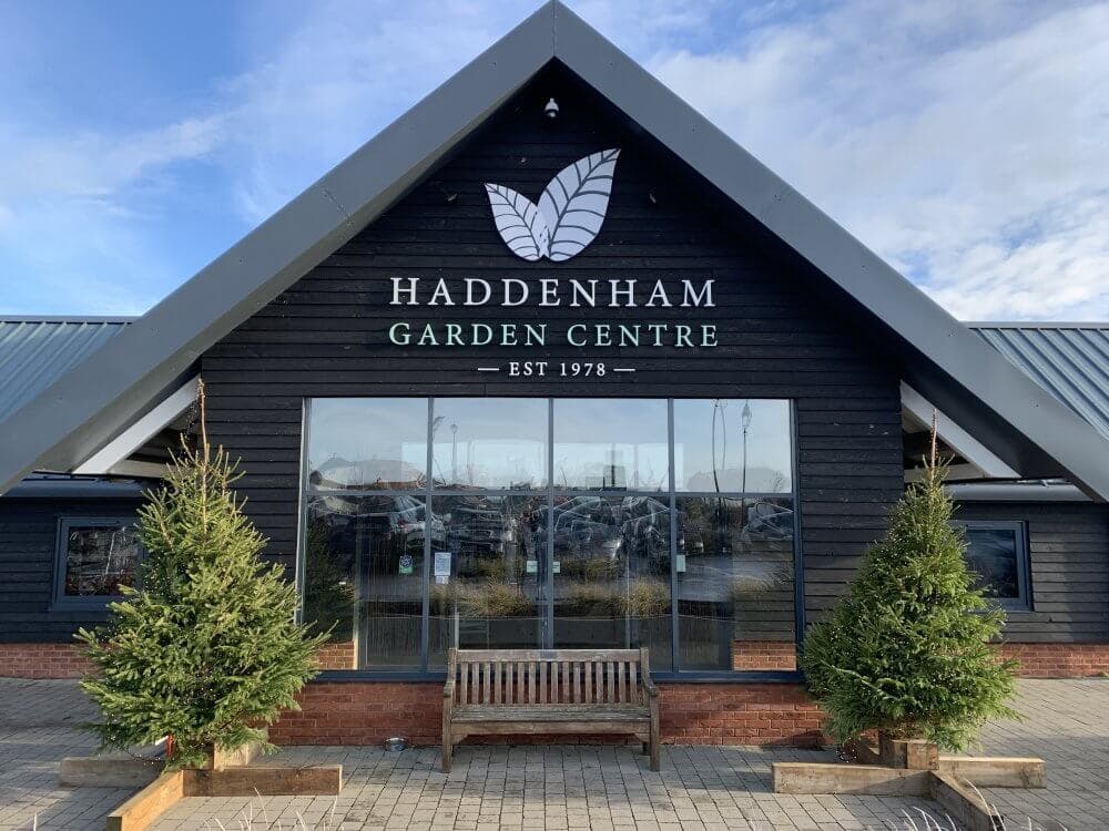 Entrance of Haddenham Garden Centre featuring a bench, two small trees, and a sign with the establishment year, 1978. - Home Instead