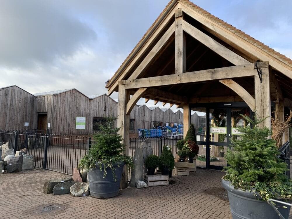 Entrance of a rustic garden center with wood-clad buildings, potted plants, and a welcoming sign on the door. - Home Instead