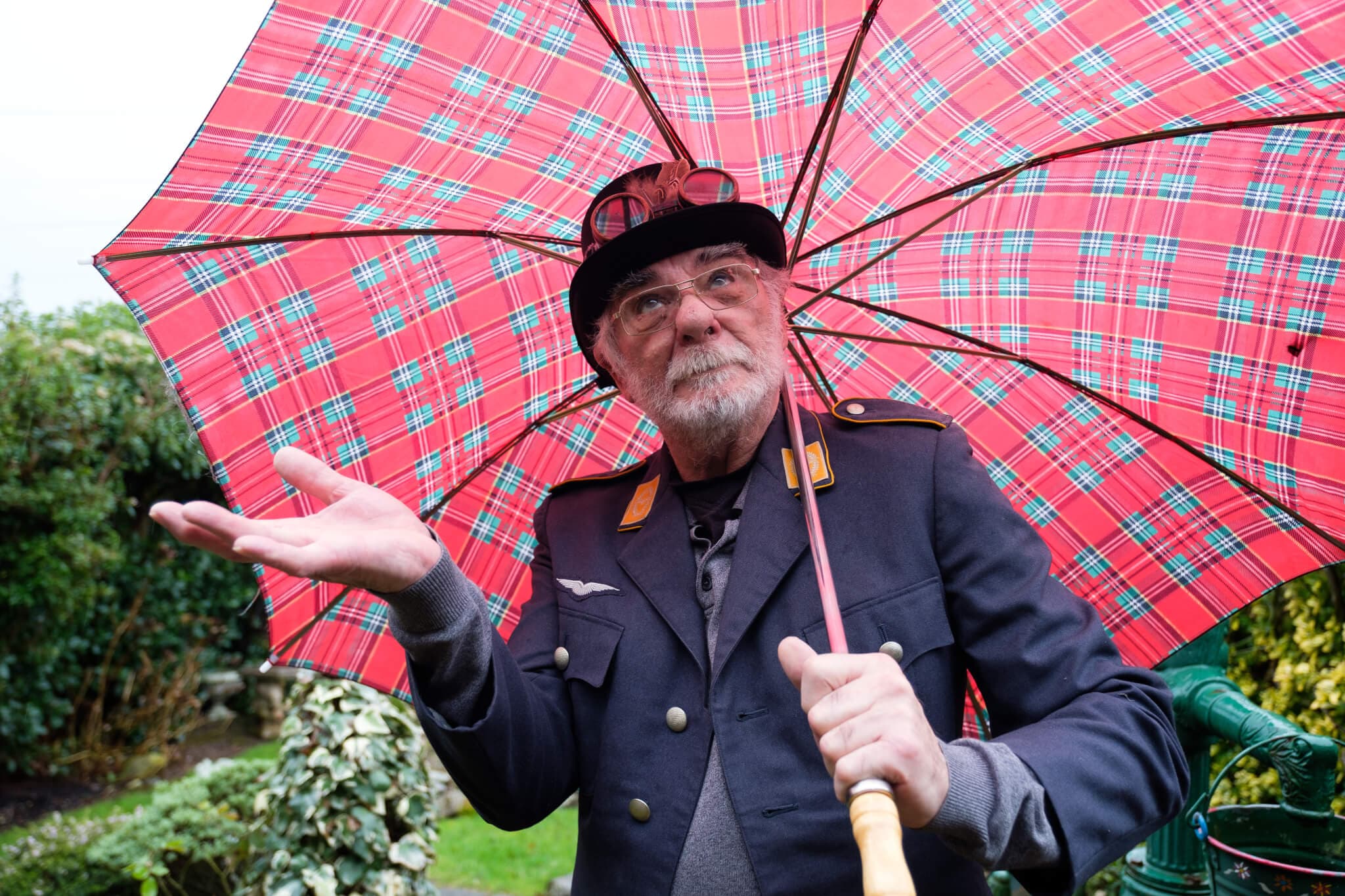 Elderly man in uniform holds a red plaid umbrella, looks up and gestures with his hand in a garden setting. - Home Instead