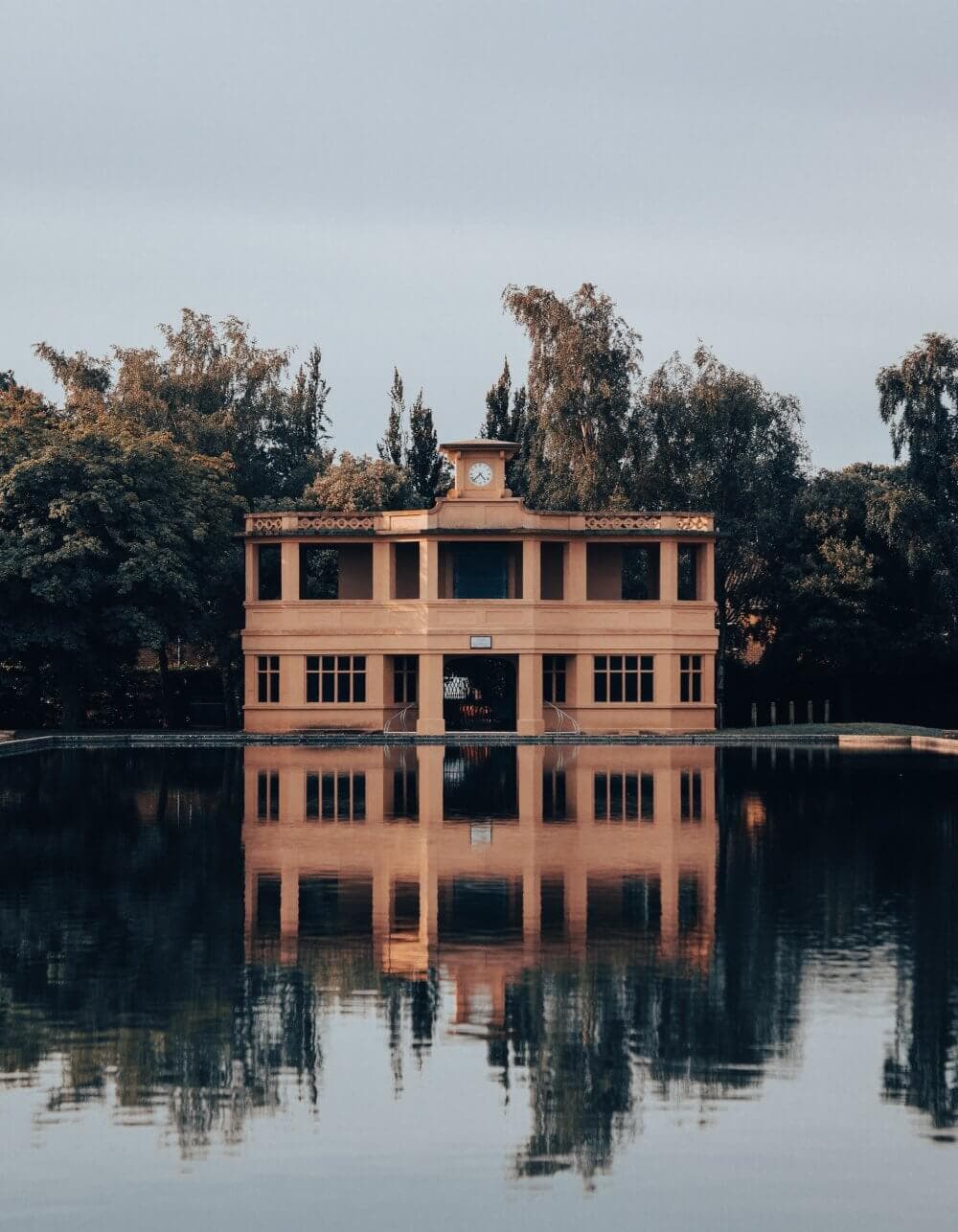Yellow building with a clock tower reflecting in a calm lake, surrounded by trees under a cloudy sky. - Home Instead