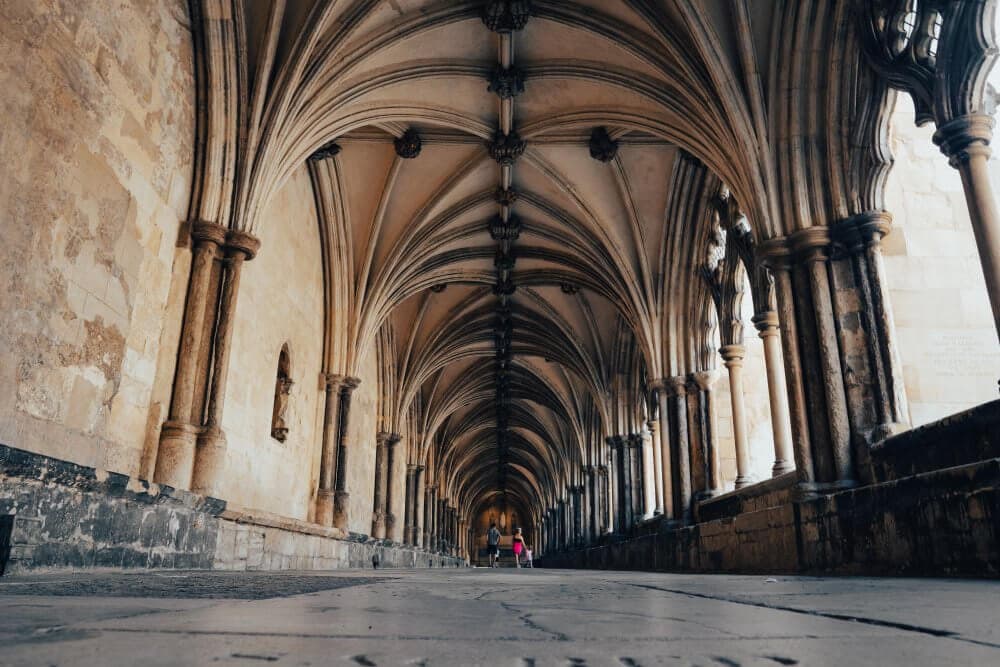 A vaulted stone corridor with gothic arches, viewed from a low angle. Two people stand in the distance. - Home Instead