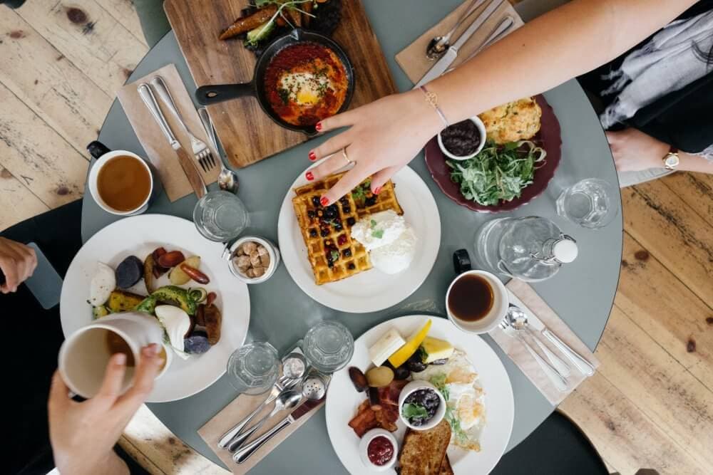 Top view of a table set with breakfast foods like waffles, eggs, toast, coffee, and hands reaching for the dishes. - Home Instead