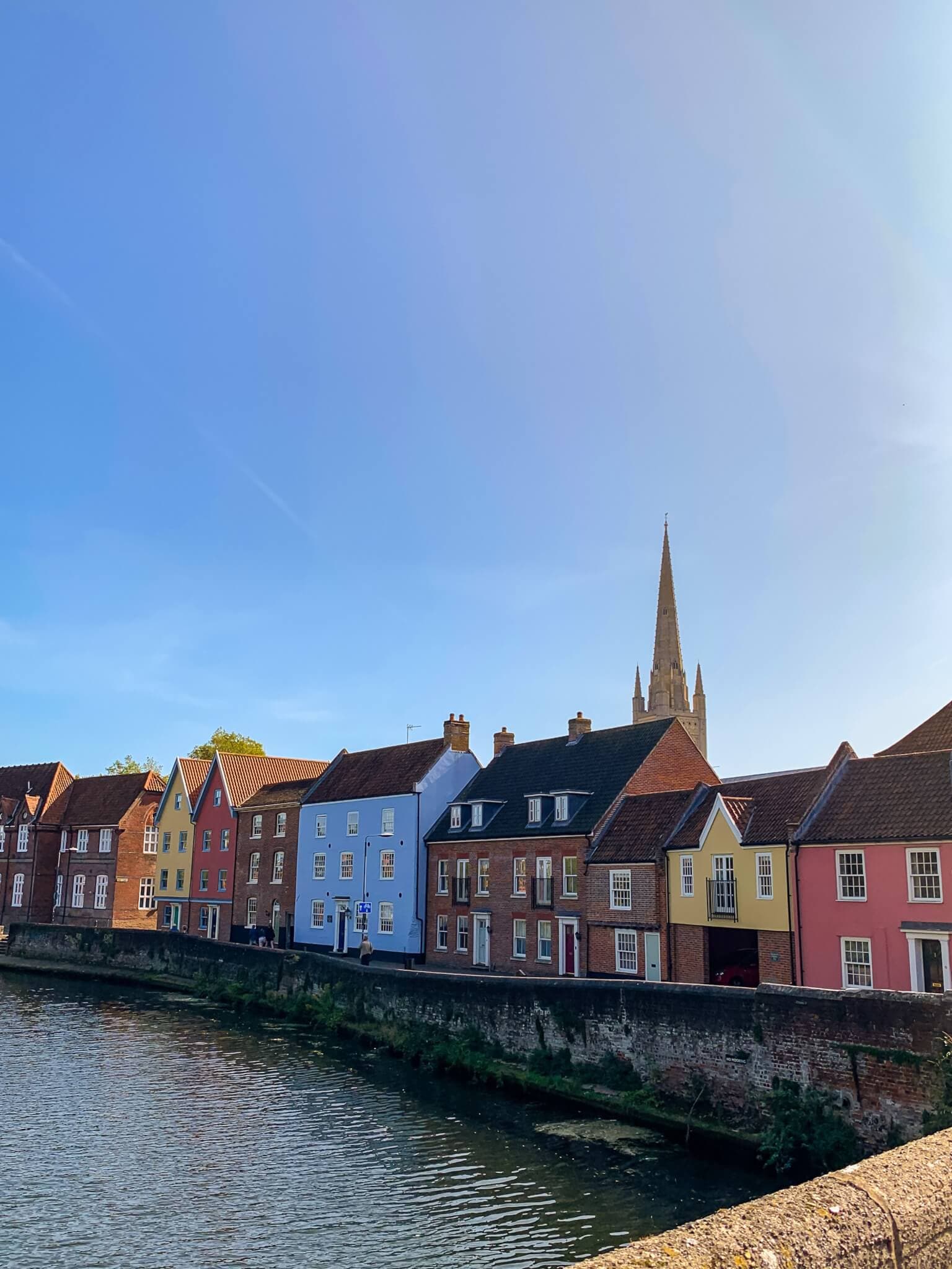 Colorful row houses along a canal with a church spire in the background under a clear blue sky. - Home Instead