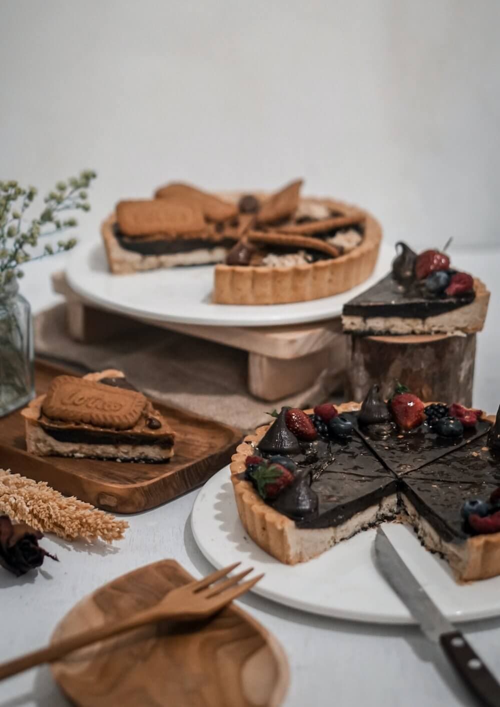 Three chocolate tarts on wooden stands, topped with assorted berries and biscuits, with a cake knife in the foreground. - Home Instead